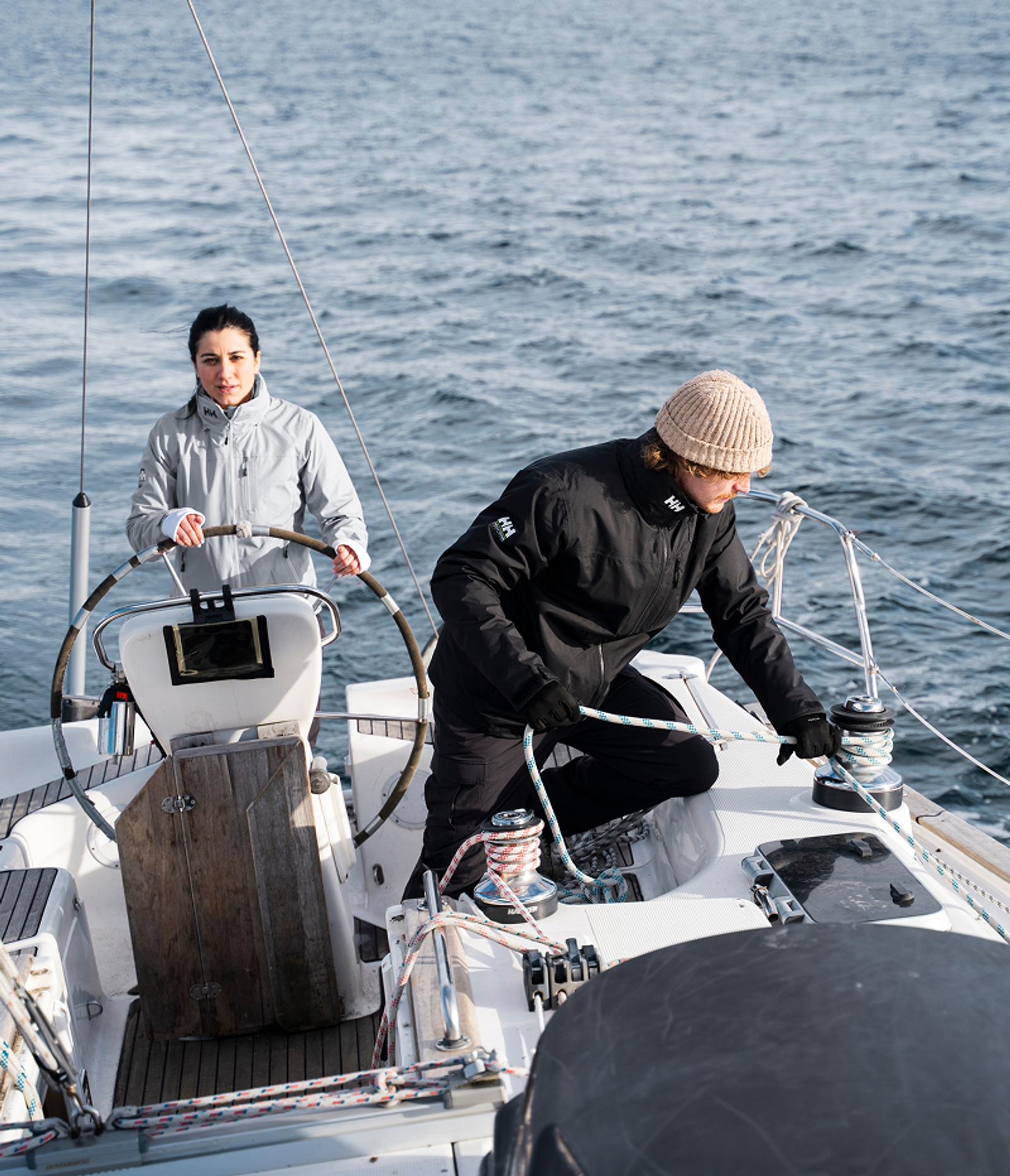 Two sailors on a boat, one steering and one working with a winch, on the open water.