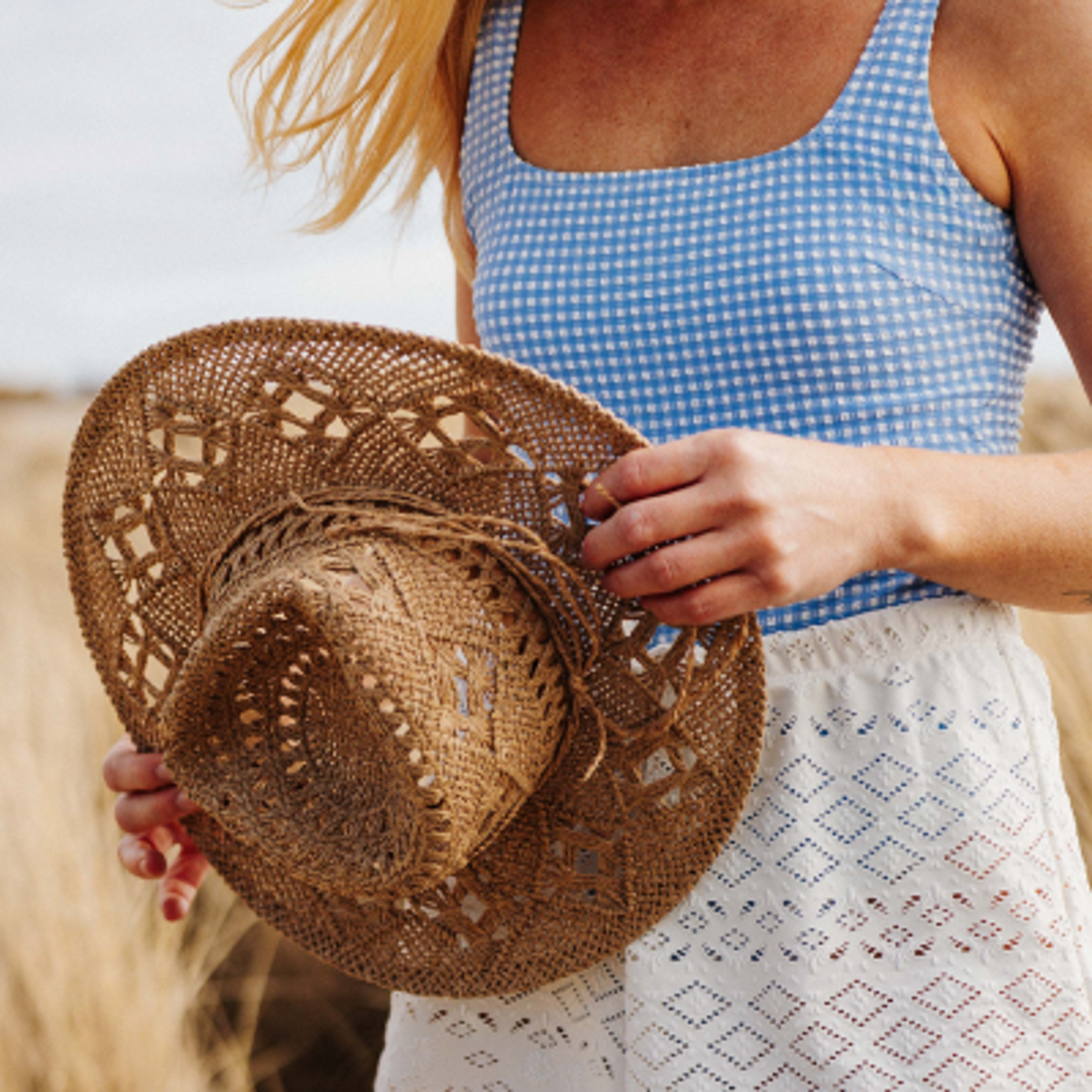 A person in a blue gingham top and white shorts holds a straw hat in a field.