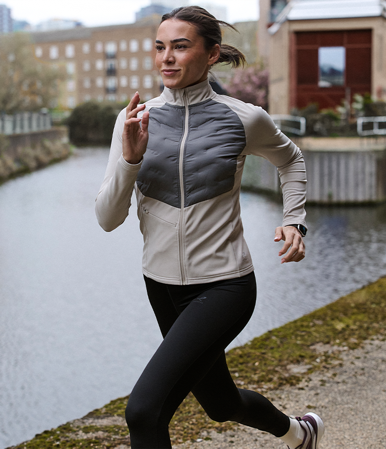 A woman in a two-tone athletic jacket and black leggings runs along a canal path.
