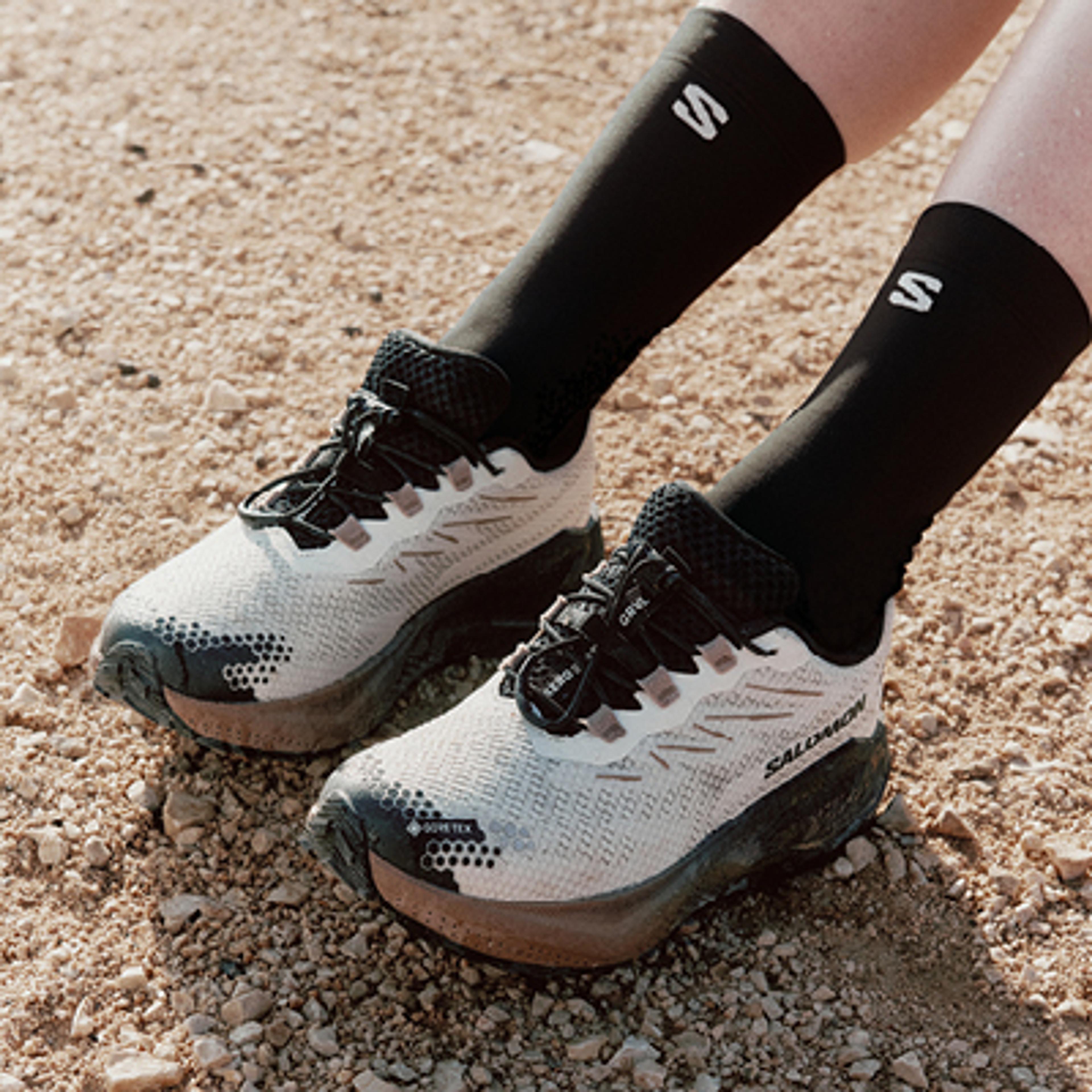 White and black Salomon Gore-Tex trail running shoes with black socks on a rocky path.