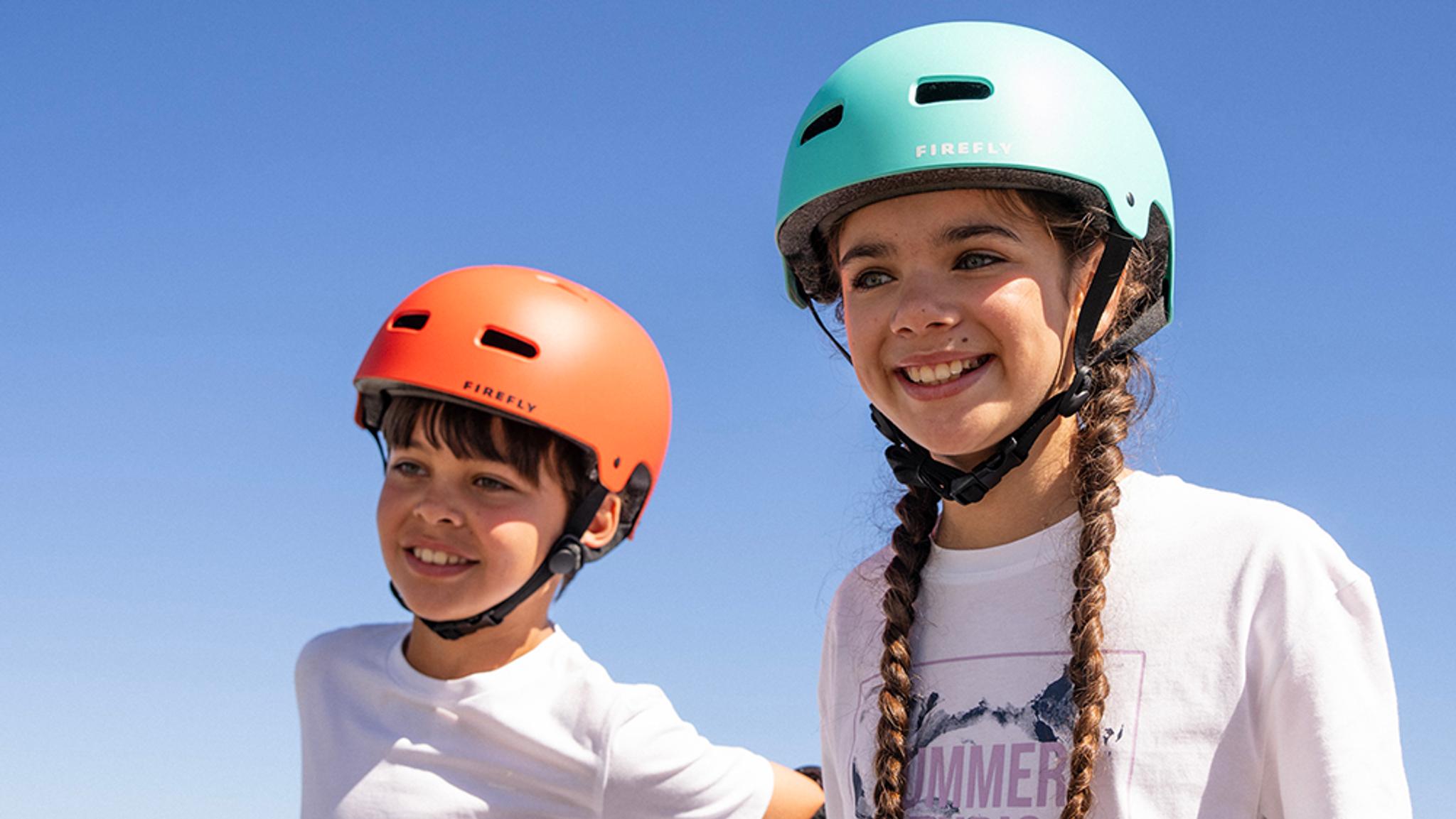 Two smiling kids, a boy and a girl, wearing orange and teal helmets against a blue sky.