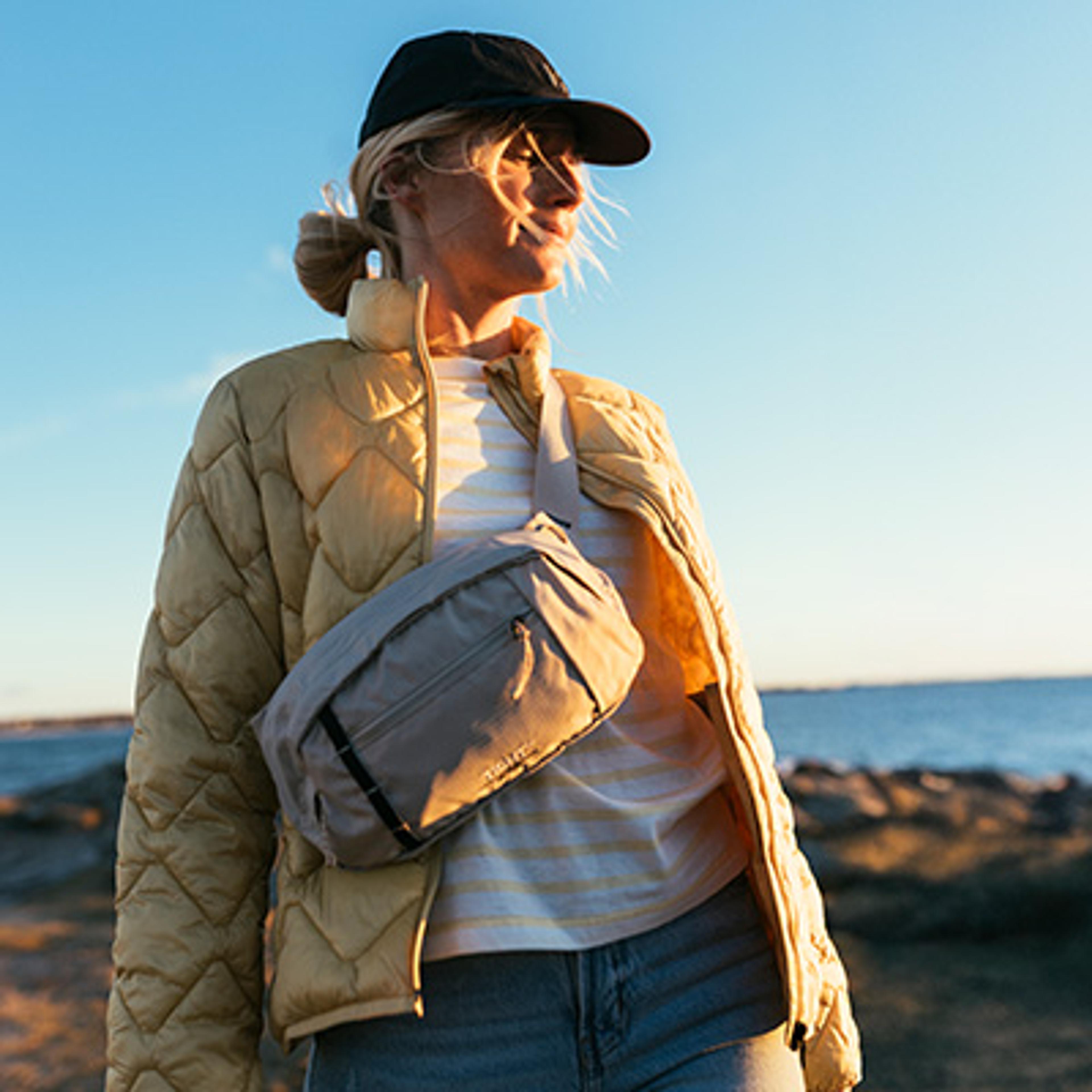 A woman in a black cap, yellow jacket, and beige sling bag looks out at the ocean at sunset.