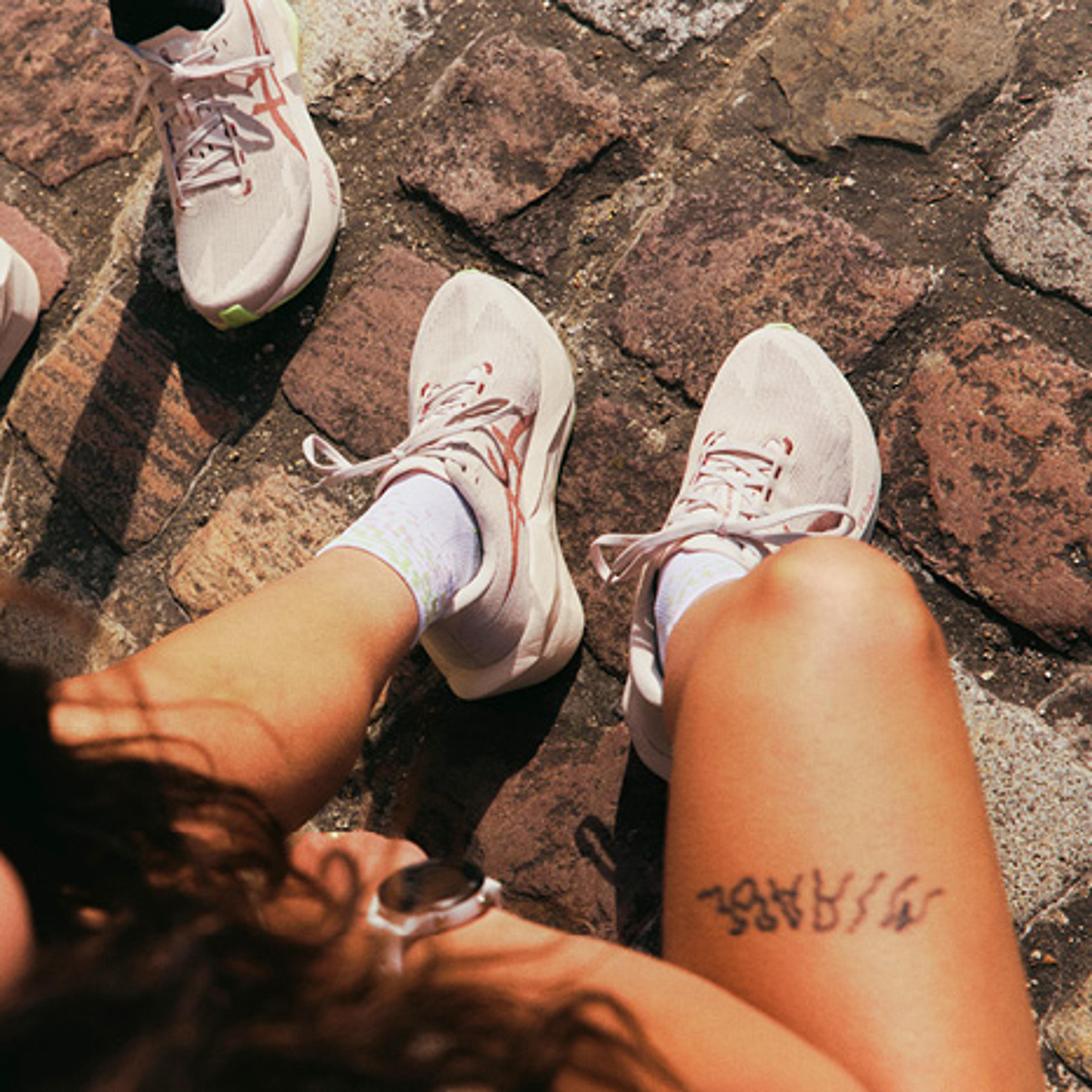 Overhead view of three people's legs in light running shoes on a cobblestone ground. One person has a tattoo on their thigh, and another's smiling face is partially visible.