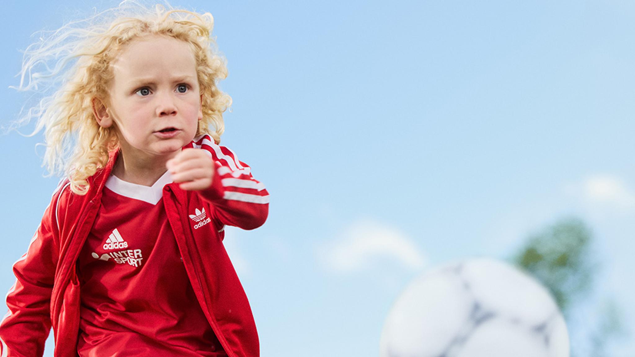 A focused child with curly blonde hair in a red soccer uniform runs under a blue sky, with a soccer ball nearby.