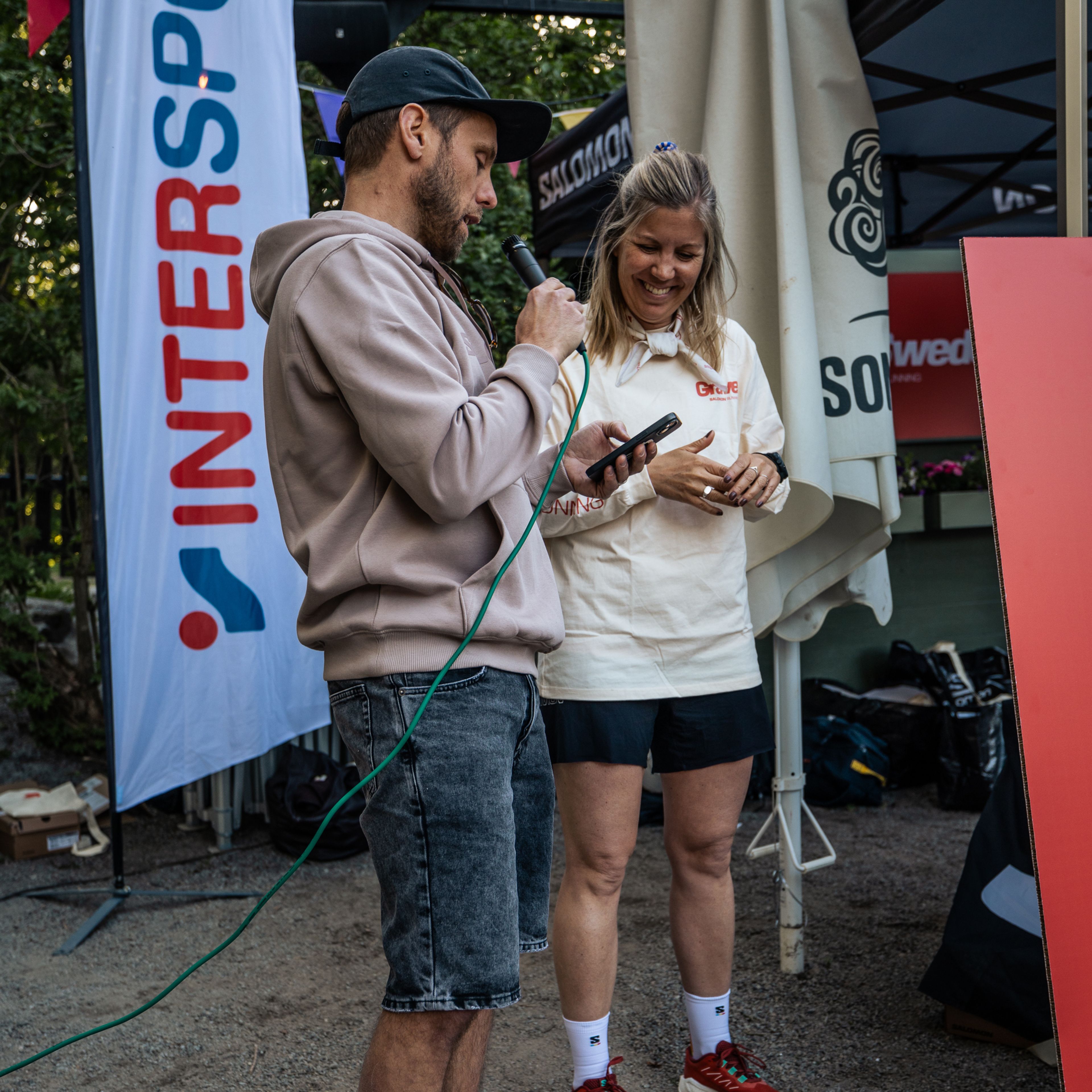 a man speaks into a microphone while a woman looks on in front of an intersport banner