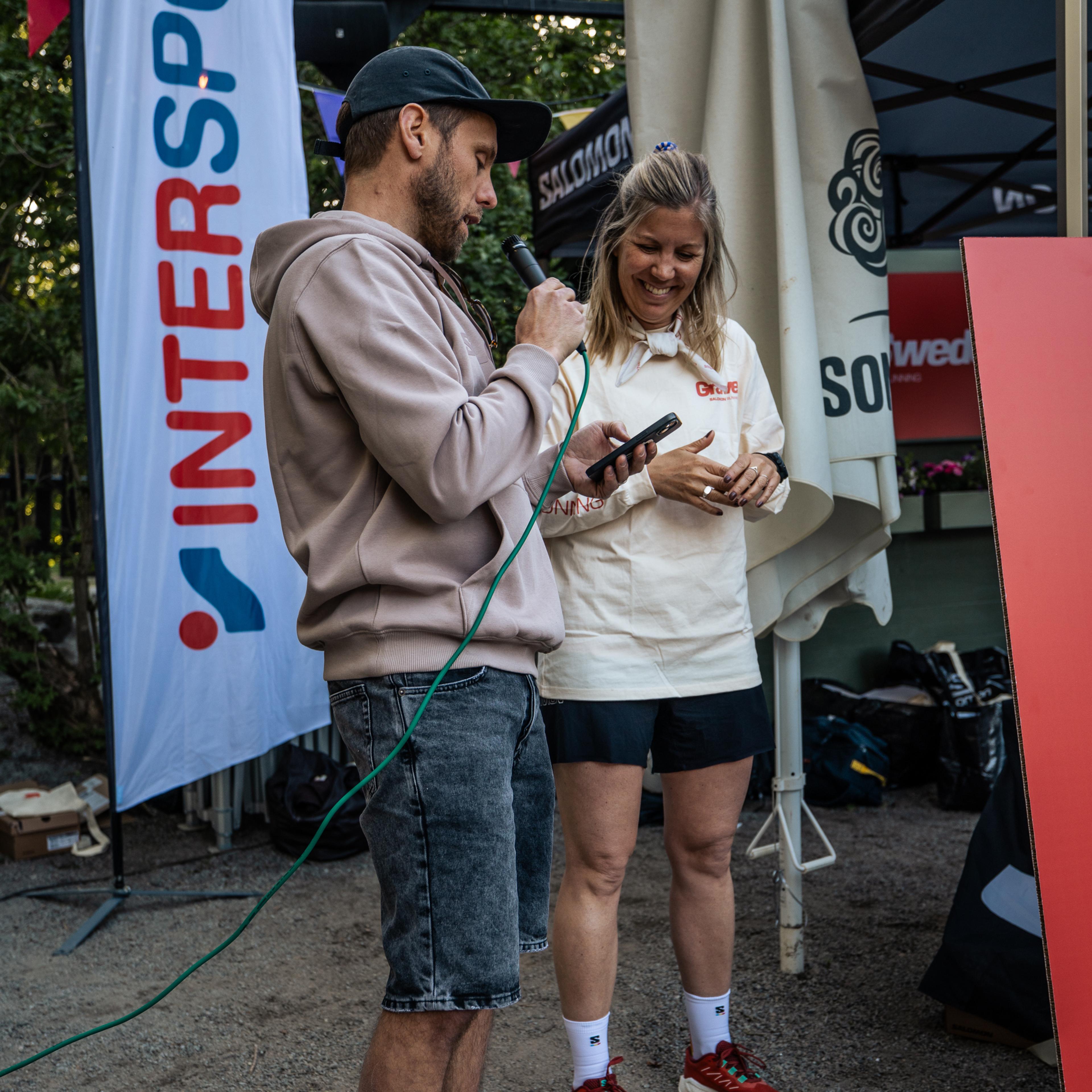 a man speaks into a microphone while a woman looks on in front of an intersport banner