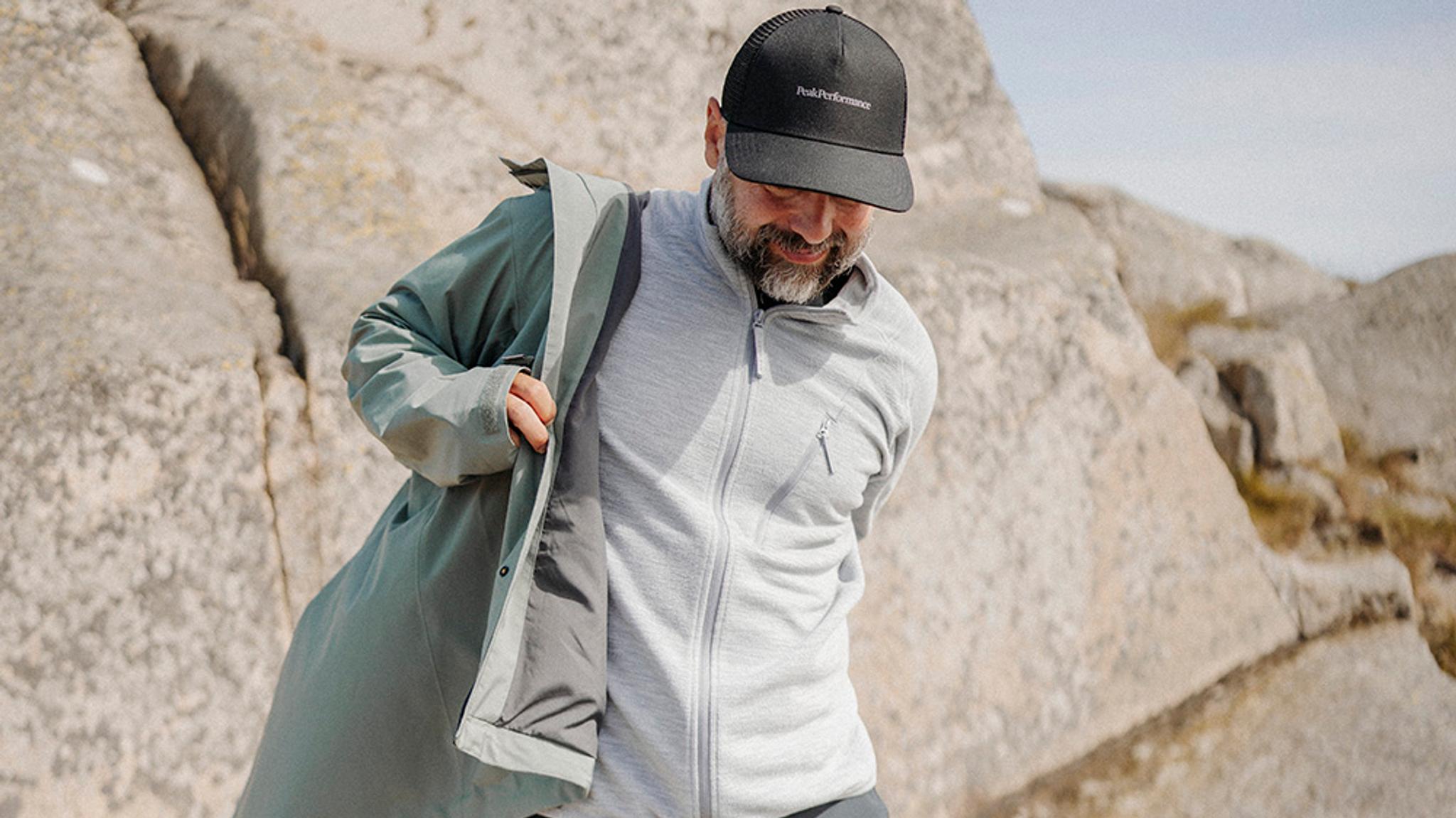 A man in a black cap and grey fleece putting on a teal jacket in a rocky outdoor setting.