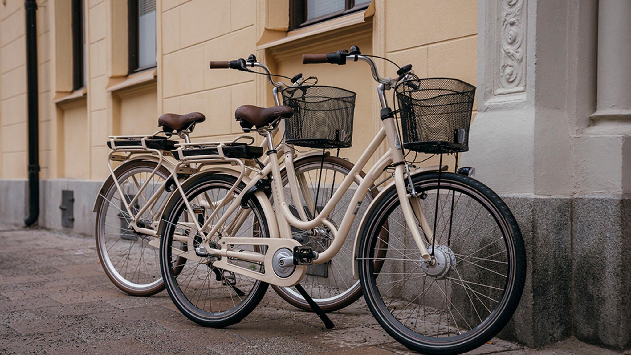 Two cream-colored bicycles with front baskets parked against a light yellow building.