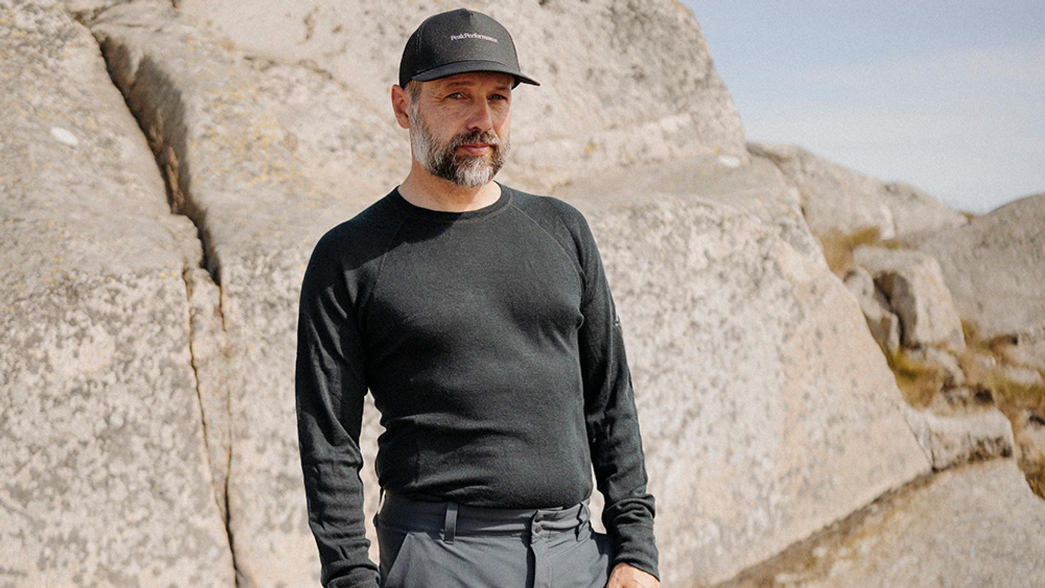 A bearded man in a black long-sleeve shirt and cap stands against a backdrop of large rocks.
