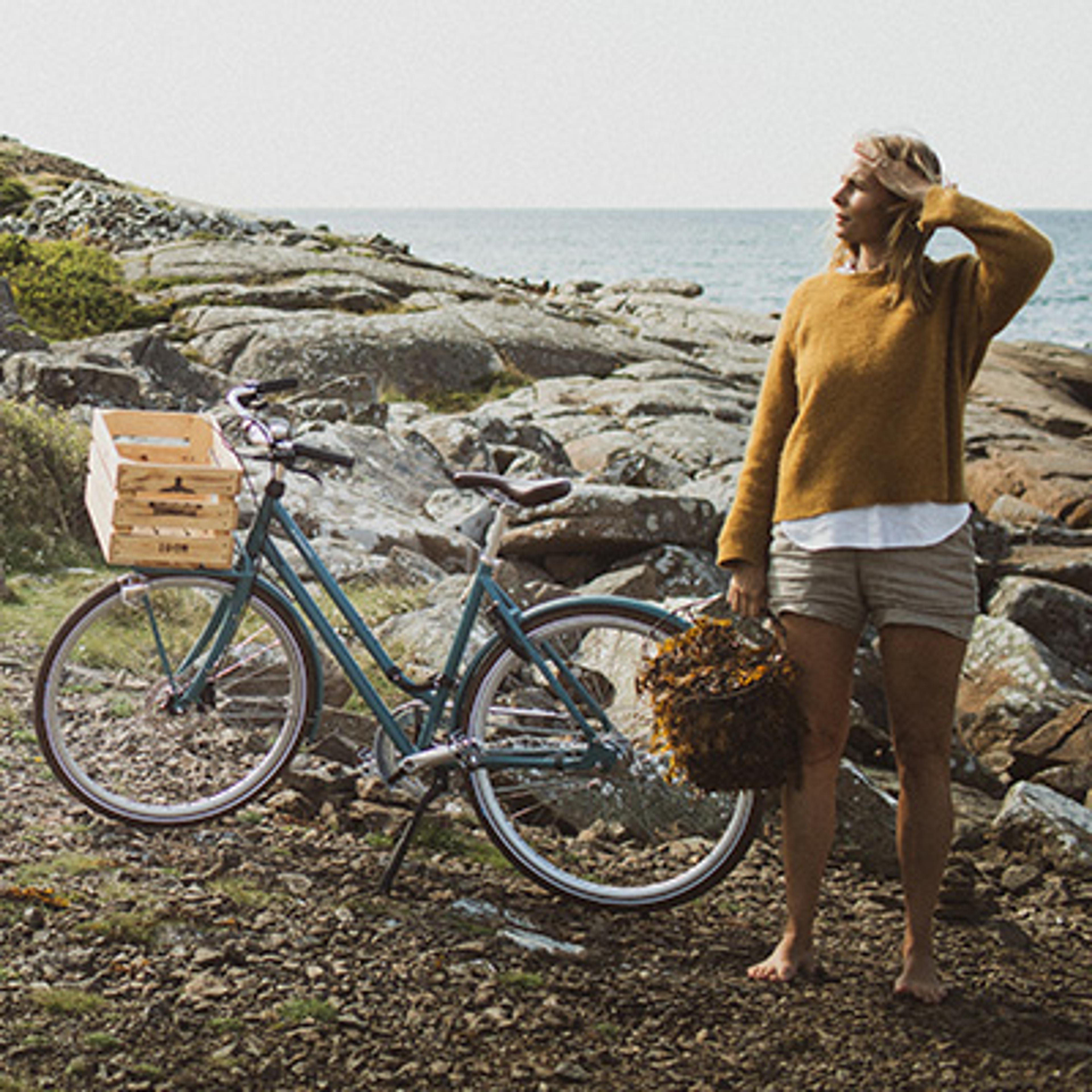 Barefoot woman holding a seaweed basket stands on a rocky shore beside a bicycle with a wooden crate, facing the ocean.