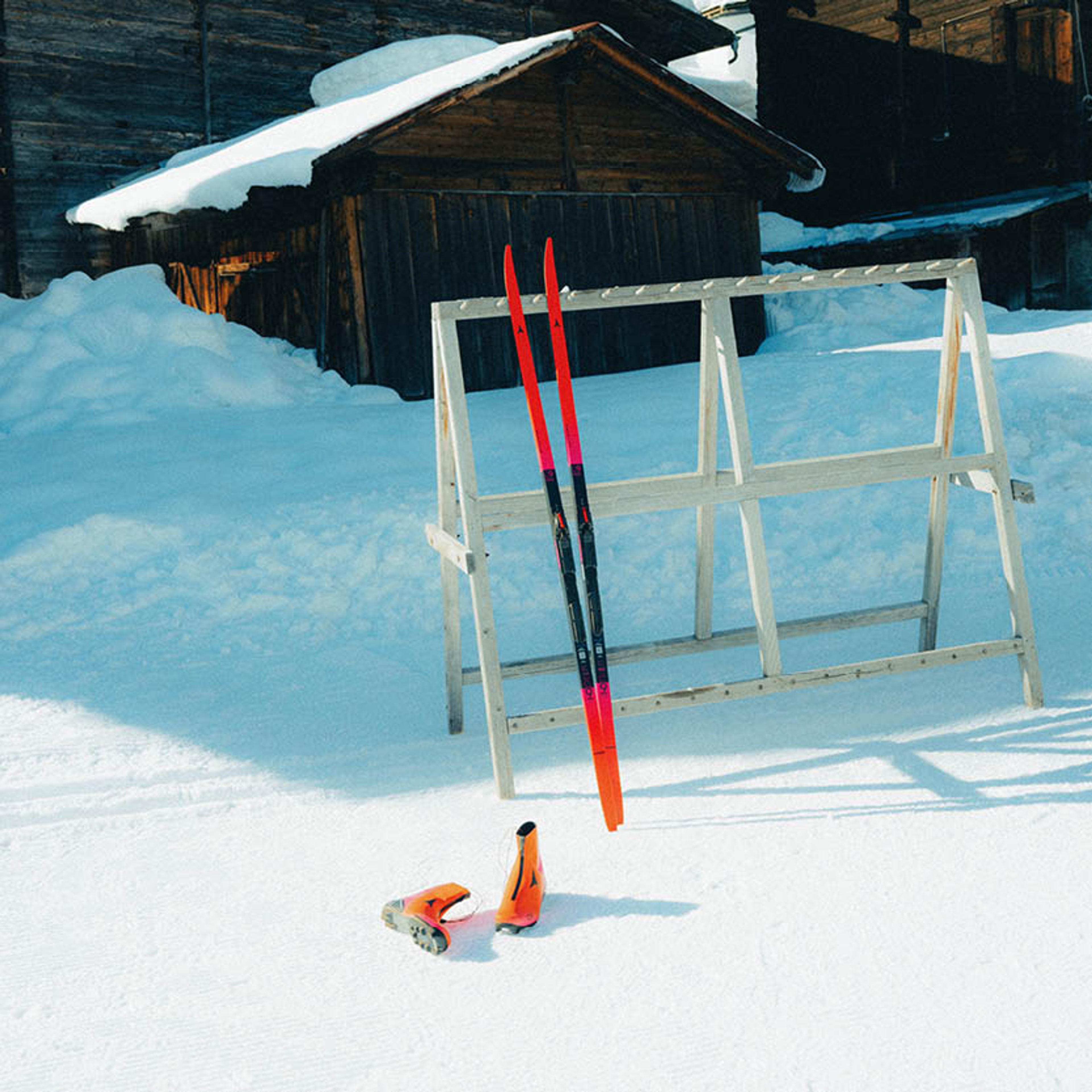 Cross-country skis and orange boots rest in fresh snow next to a wooden ski rack, with snowy buildings in the background.