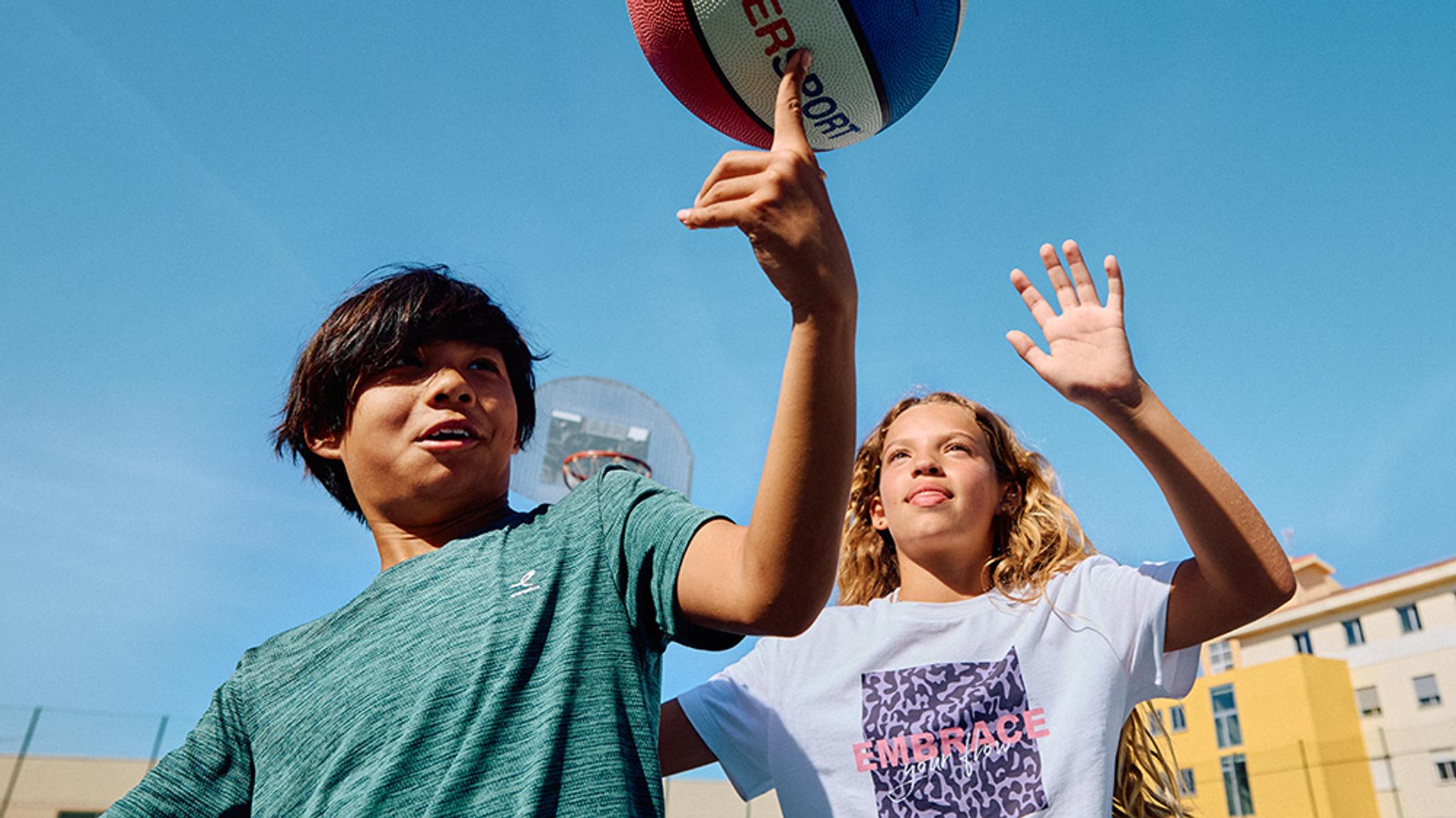 A boy spins a basketball on his finger while a girl stands with her hand raised, outdoors under a blue sky.