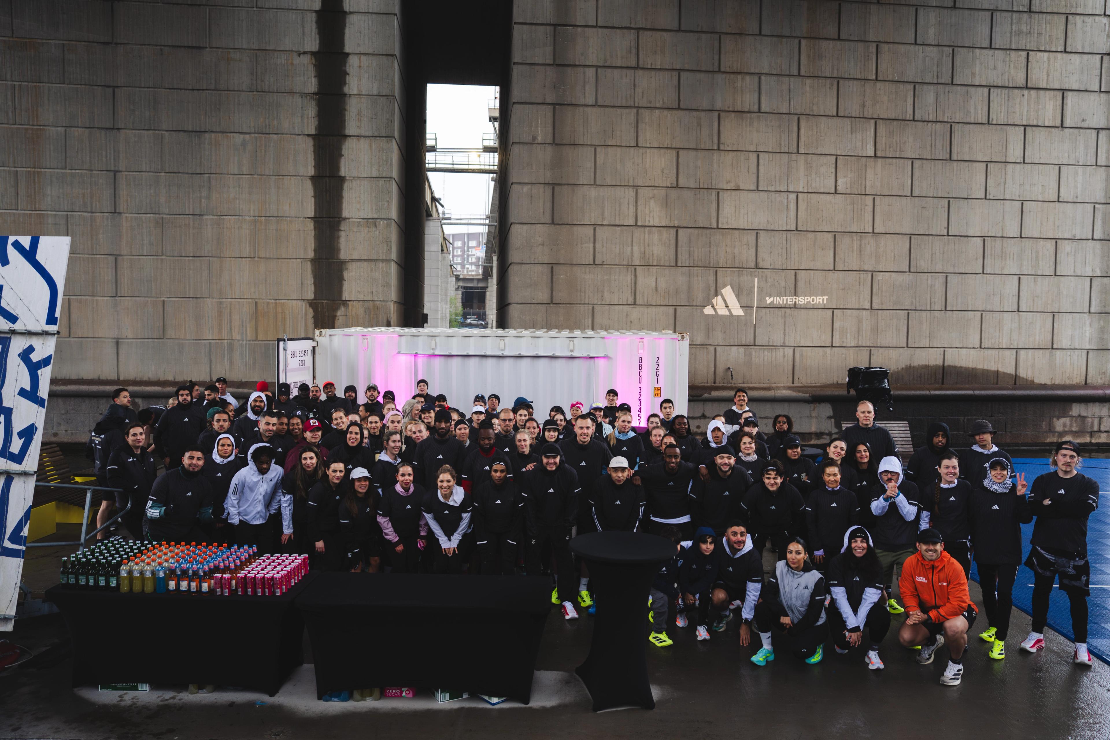 A large group of people in athletic wear pose outdoors in front of a concrete wall with an Adidas logo, shipping containers, and tables of colorful drinks.