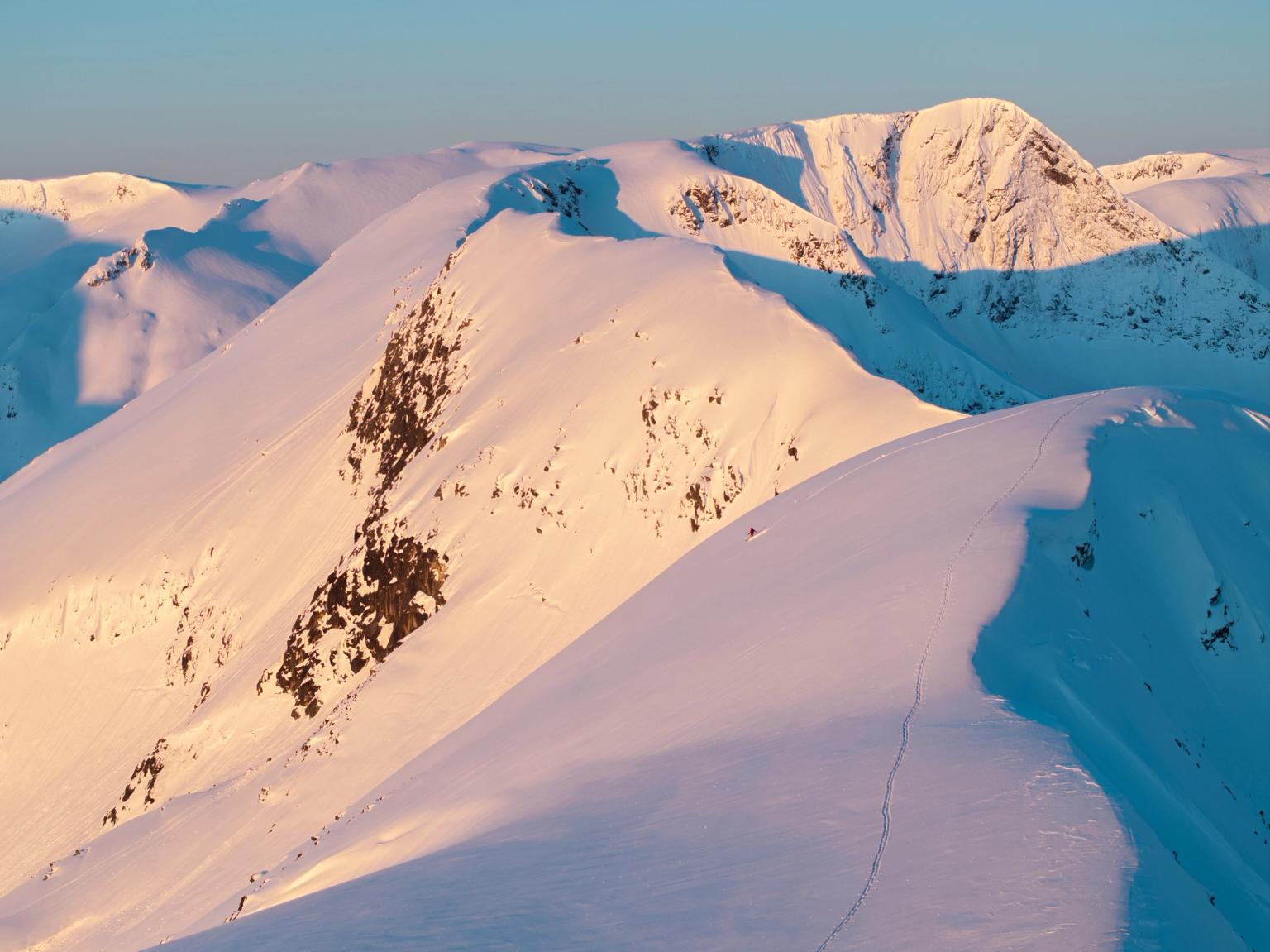 A skier descends a snow-covered mountain ridge, bathed in golden light and deep blue shadows.