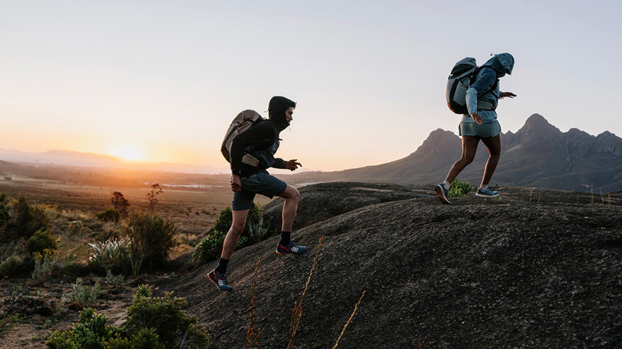 Two hikers with backpacks ascend a rocky hill at sunset.