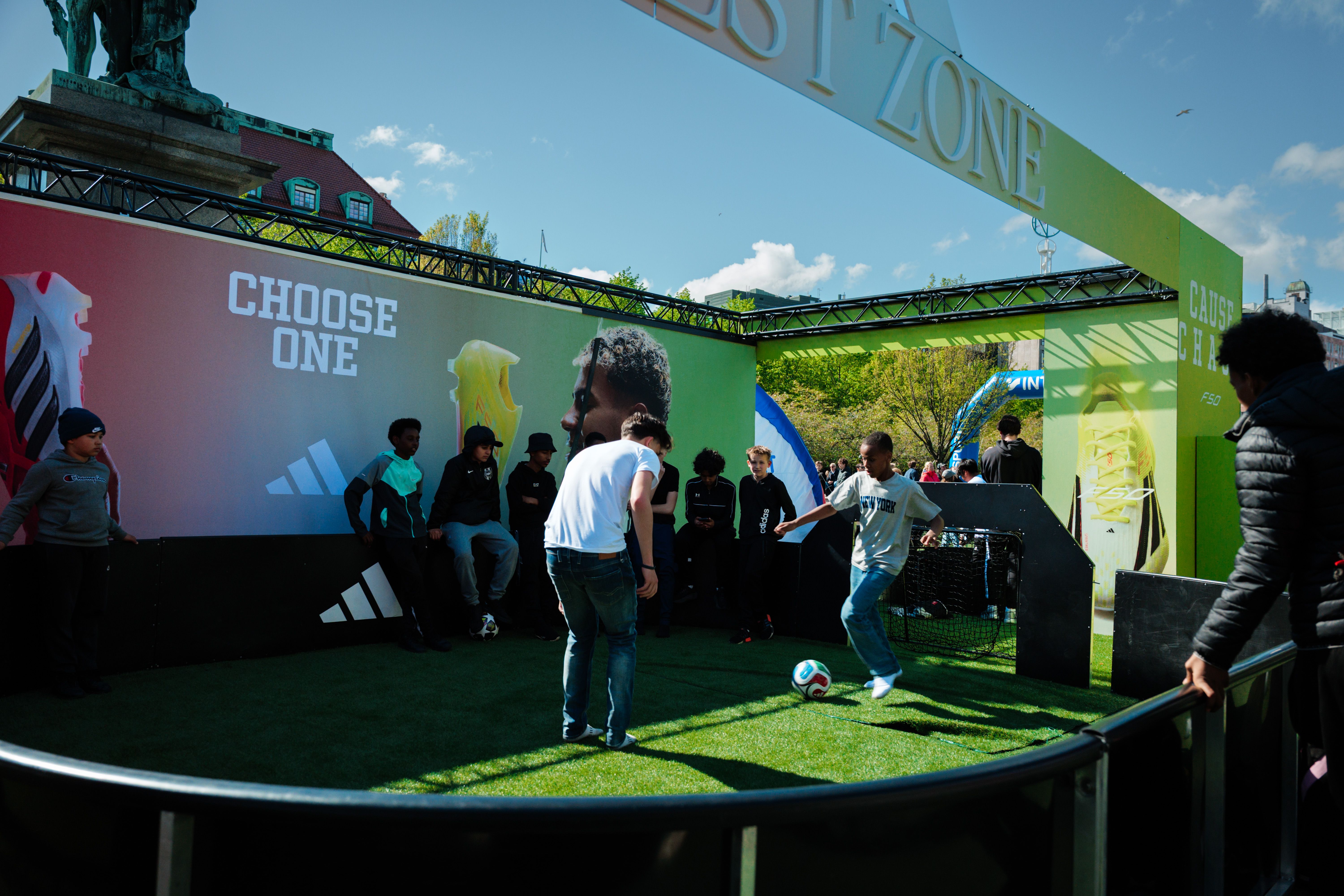 Young people play soccer on artificial turf at an outdoor Adidas "TEST ZONE" event, with a "CHOOSE ONE" banner in the background.