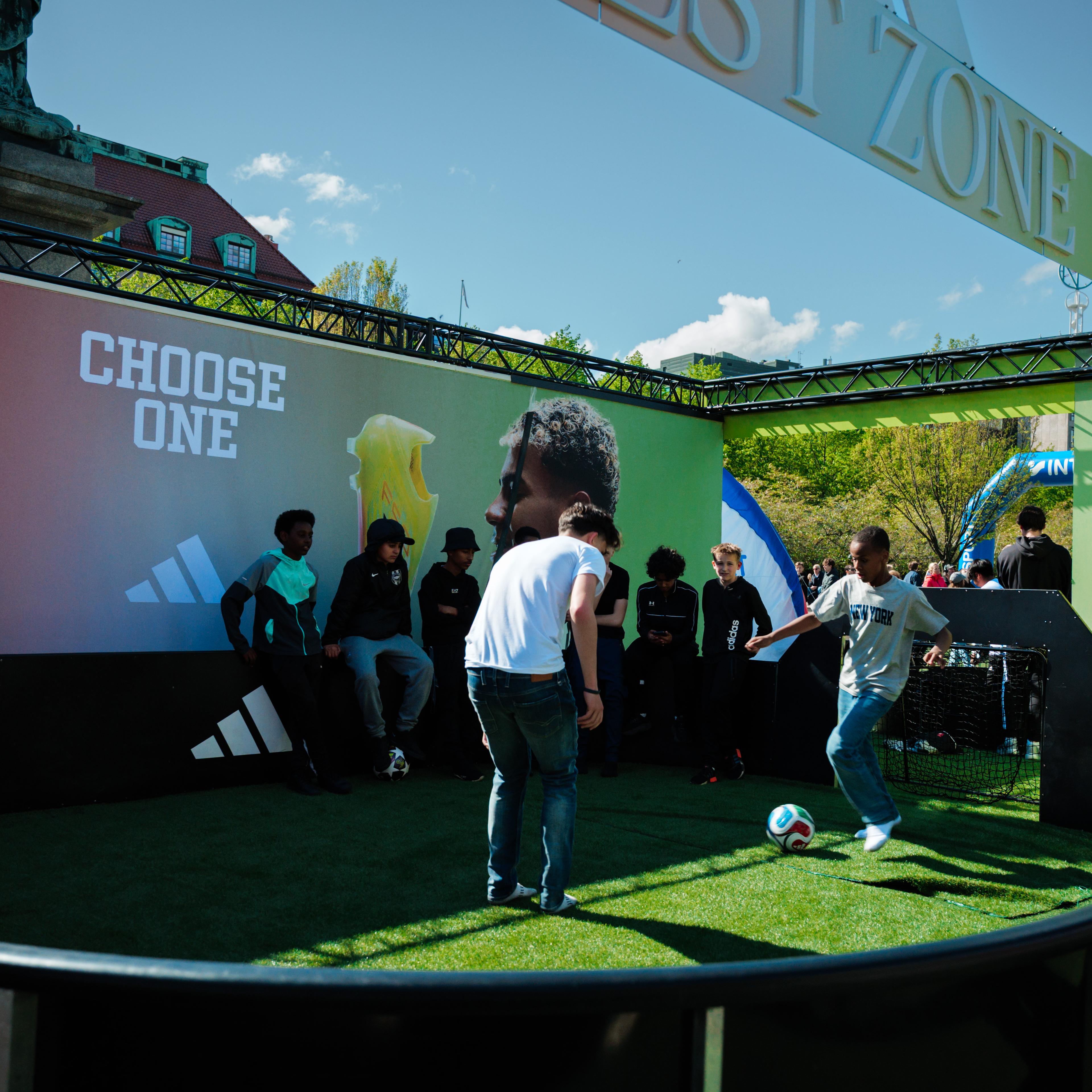 Young people play soccer on artificial turf at an outdoor Adidas "TEST ZONE" event, with a "CHOOSE ONE" banner in the background.