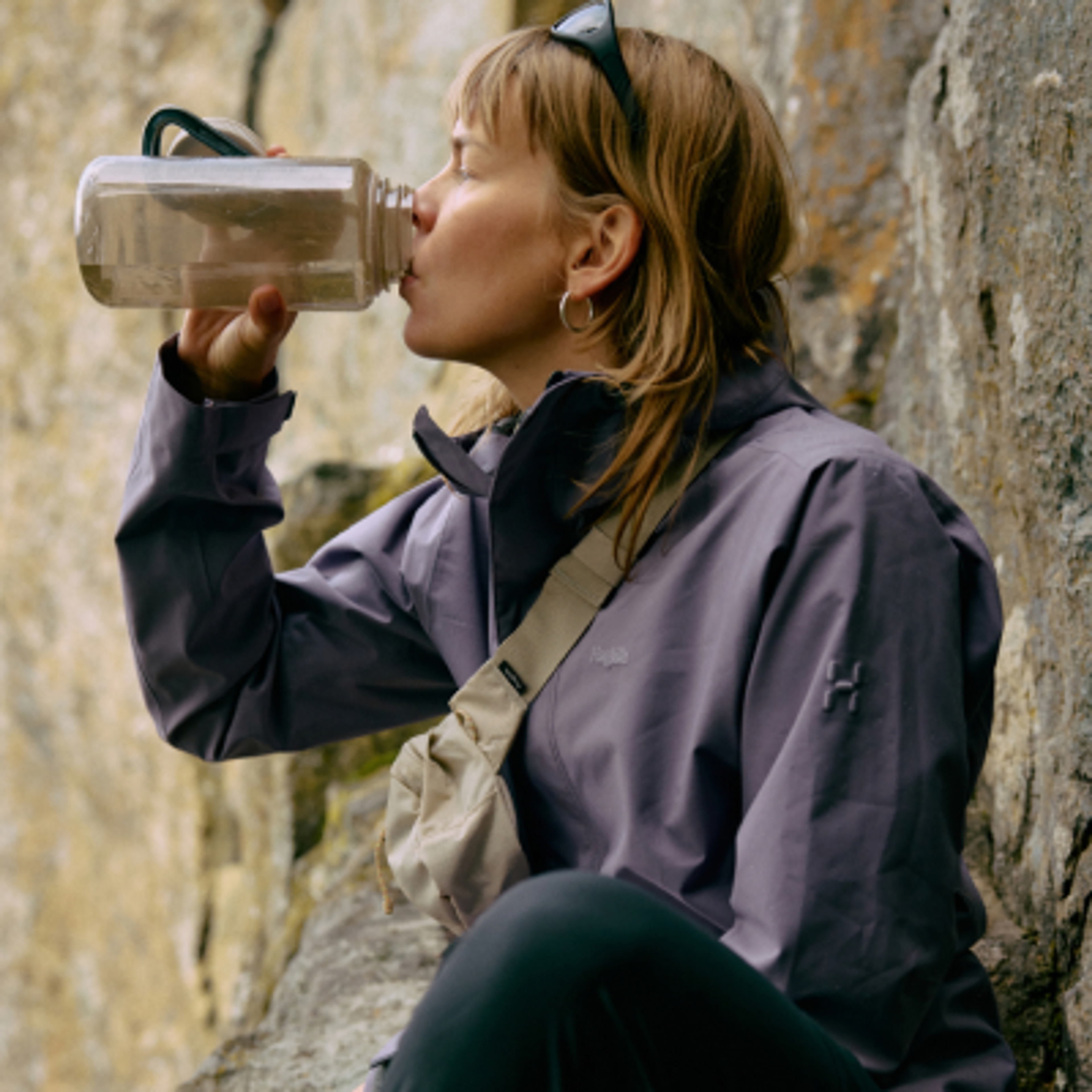 A woman in a purple jacket drinks from a water bottle against a rocky background.