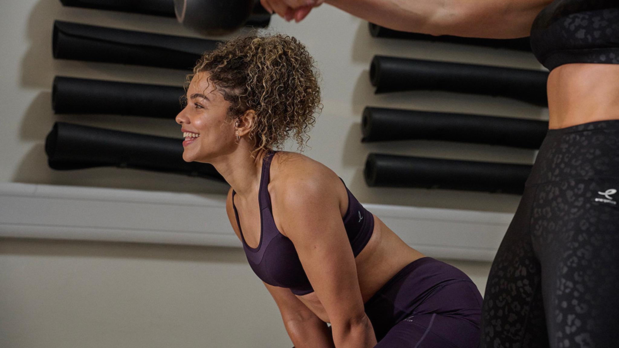 A smiling woman in purple activewear exercises alongside another woman in a gym.