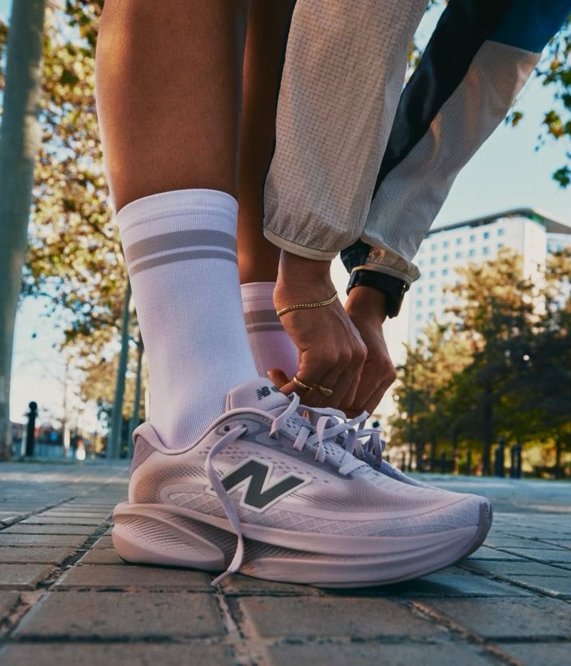 A person tying the laces of a light purple New Balance running shoe on a paved path.