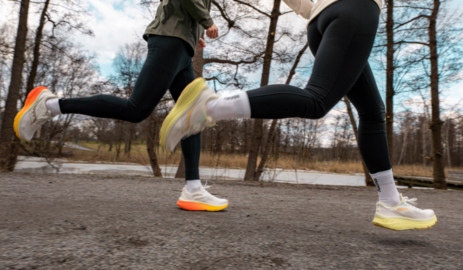 Two runners' legs in black leggings and white shoes with bright soles mid-stride on a dirt path.