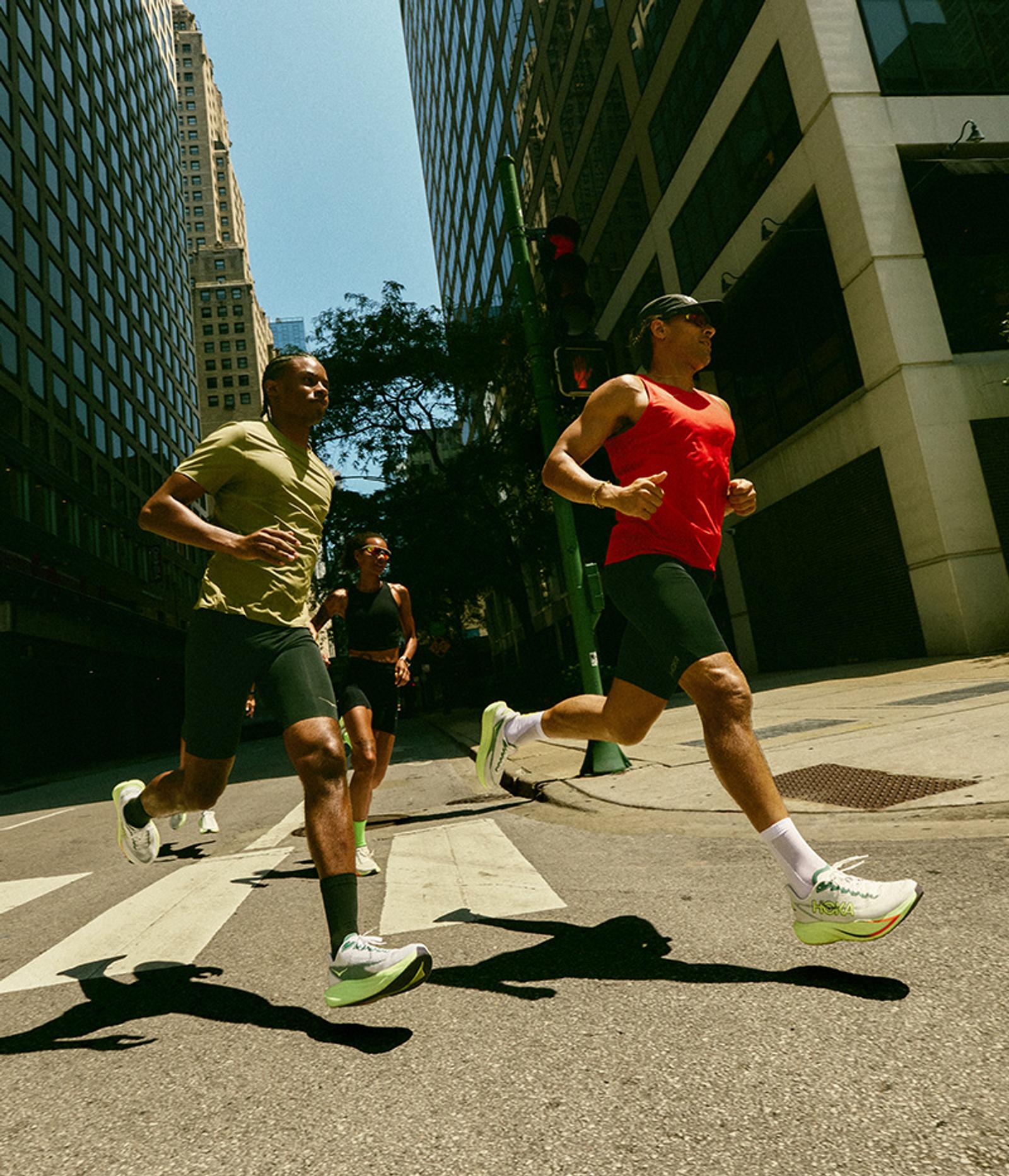 Three athletes run across a city street with tall buildings in the background.