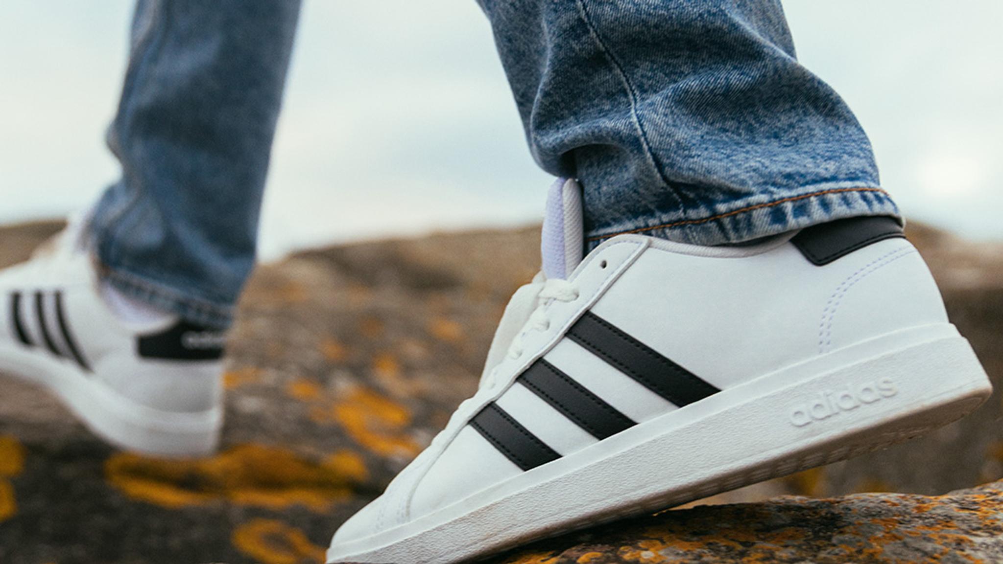 Person in blue jeans and white Adidas sneakers with black stripes walking on rocky ground.