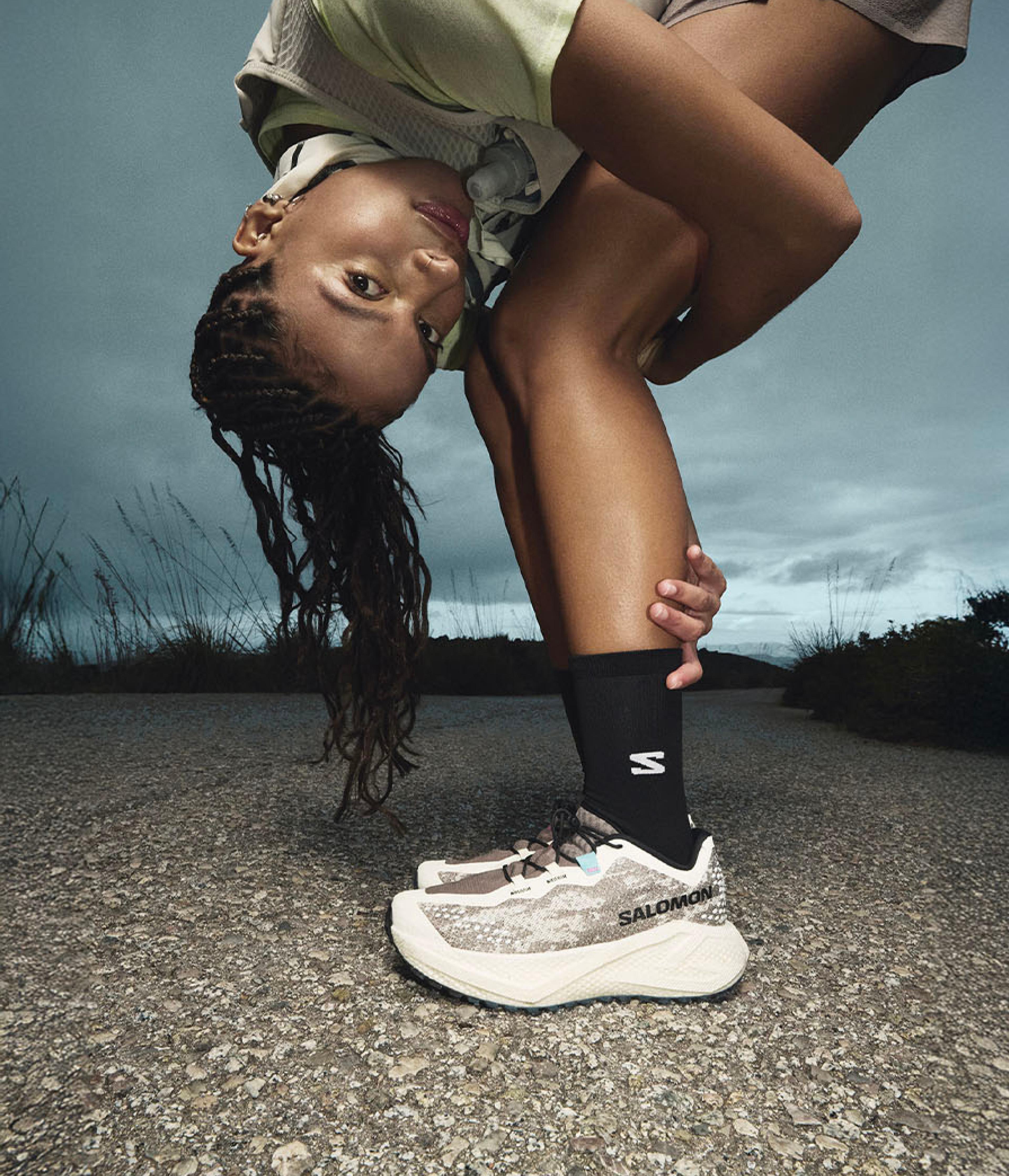 A woman in Salomon trail running shoes bends to stretch, looking at the camera, on a road under a dark sky.