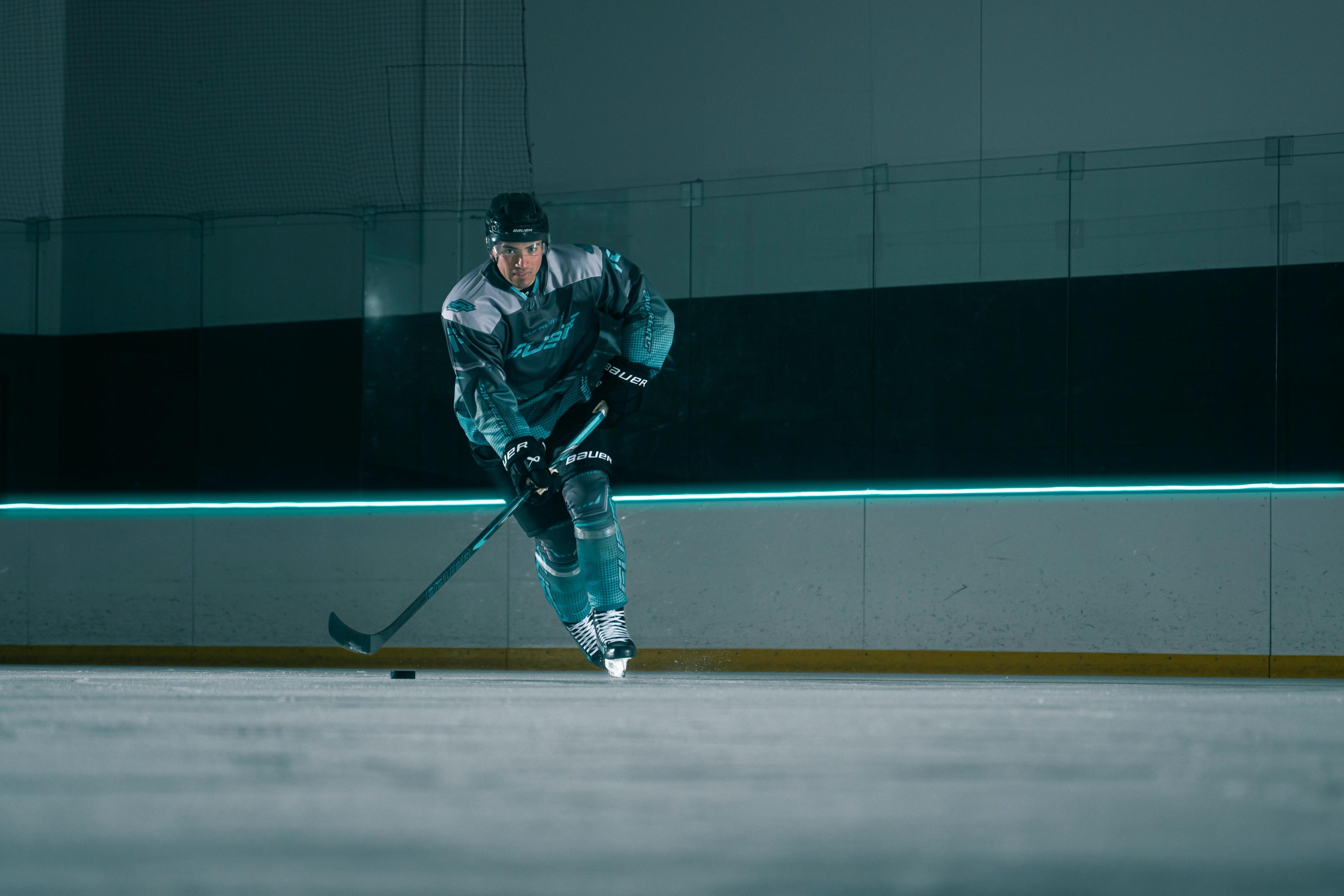 A hockey player in a teal uniform skates on an ice rink, dribbling a puck, with a neon teal light strip in the background.