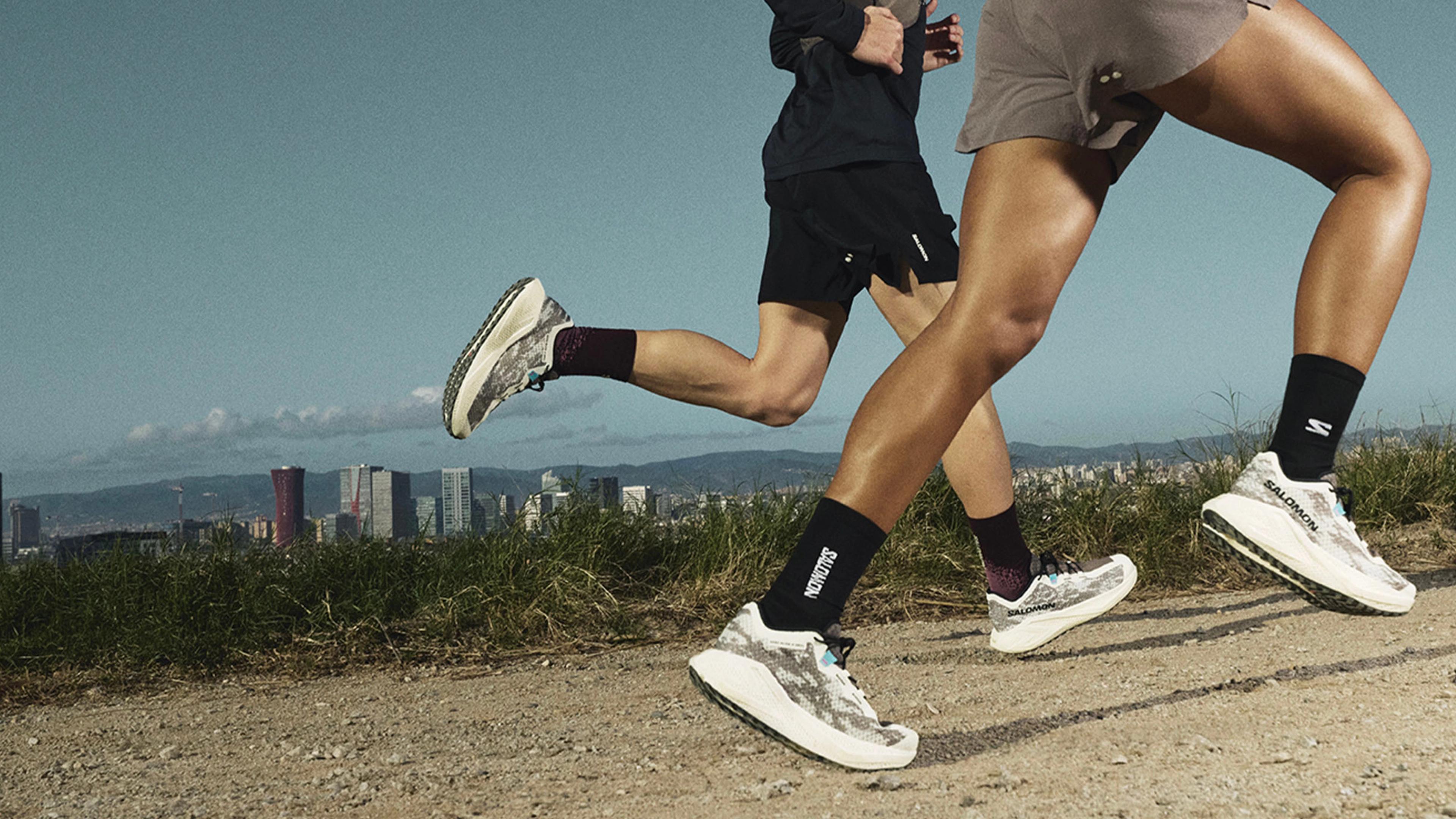 Two people run in Salomon shoes on a dirt path, with a city skyline in the background.