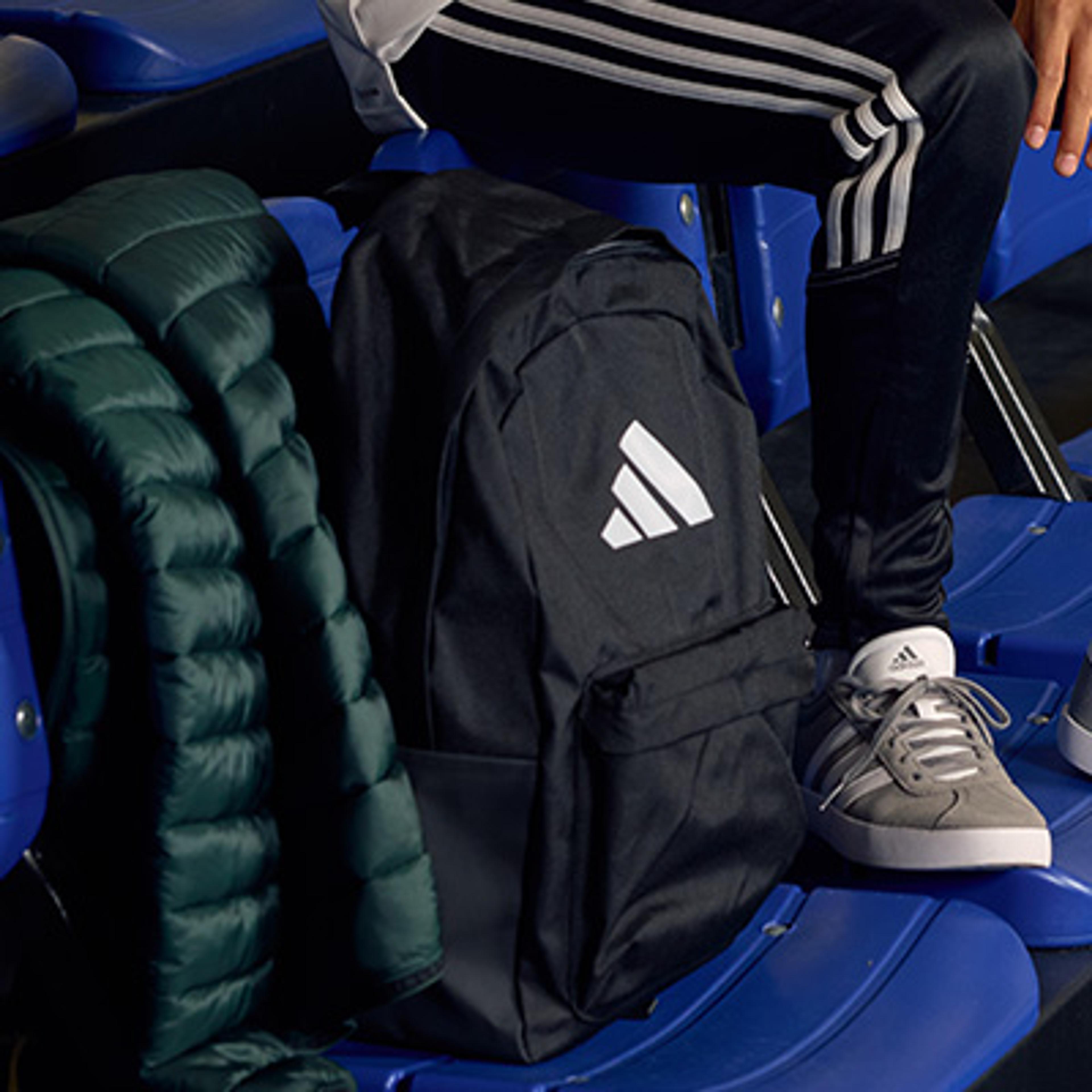 A person wearing Adidas track pants and shoes sits next to an Adidas backpack and a green puffer jacket on blue stadium seats.