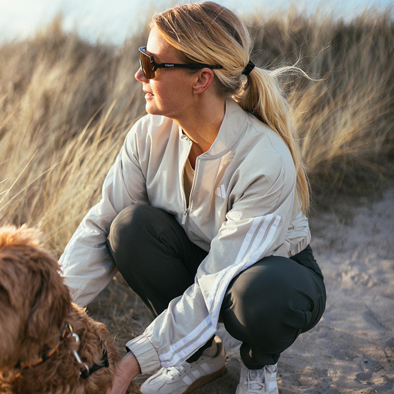 Woman in sunglasses and athletic wear crouching to pet a dog on a sandy dune with tall grass.