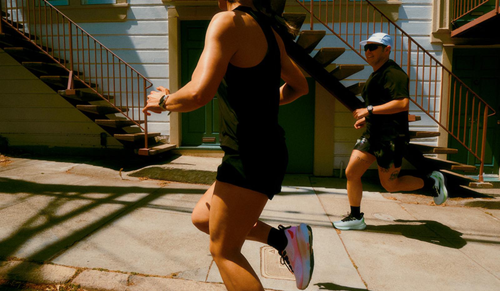 Two people running down a sunny urban street past an apartment building with outdoor stairs.