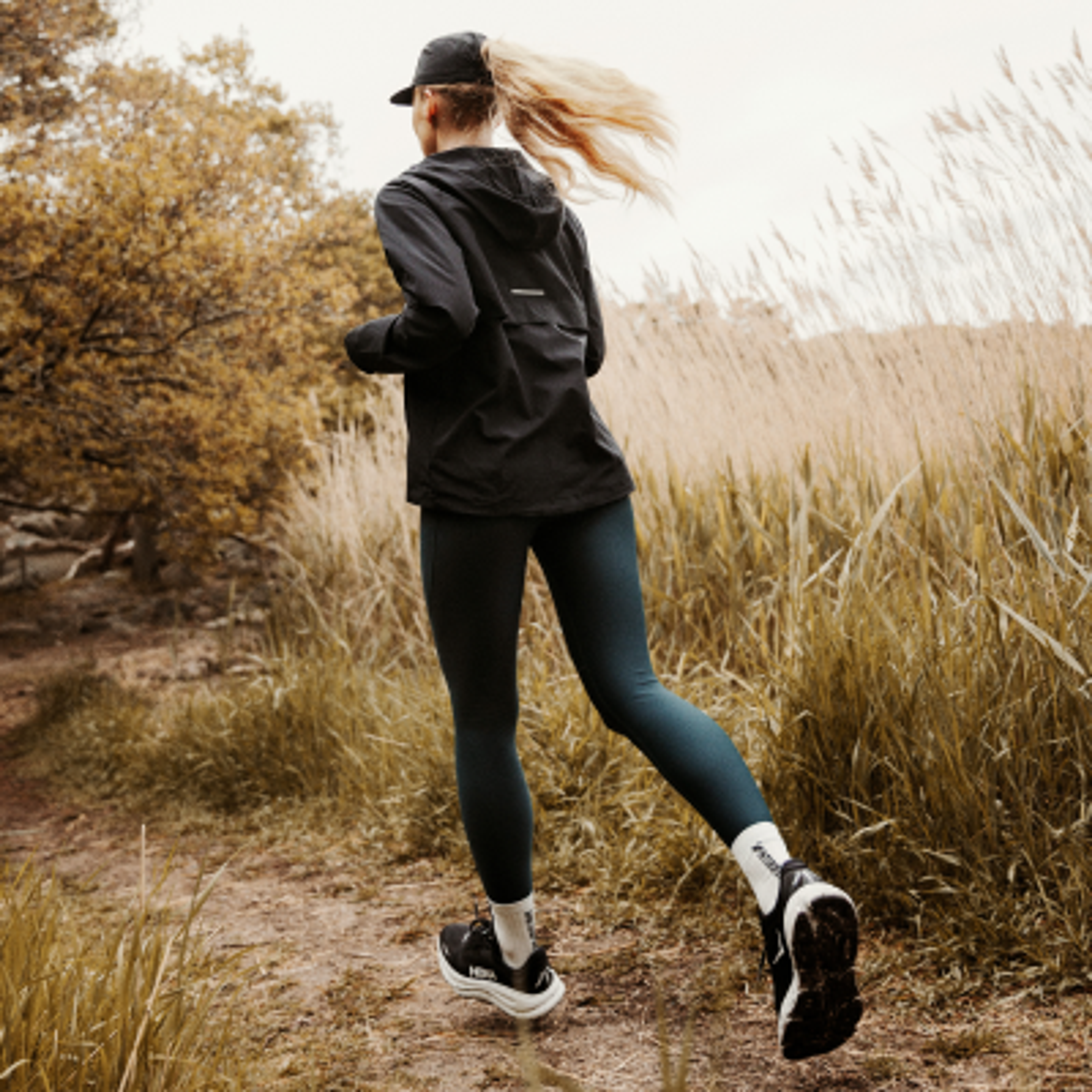 A woman in a black jacket and dark leggings runs on a dirt path through tall golden grass, viewed from behind.