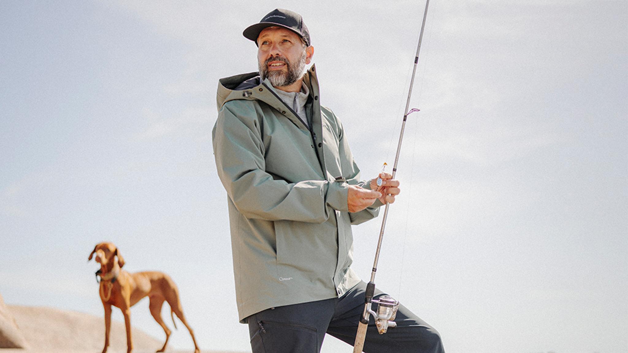 A man in a green jacket and cap holds a fishing rod, with a brown dog in the background.