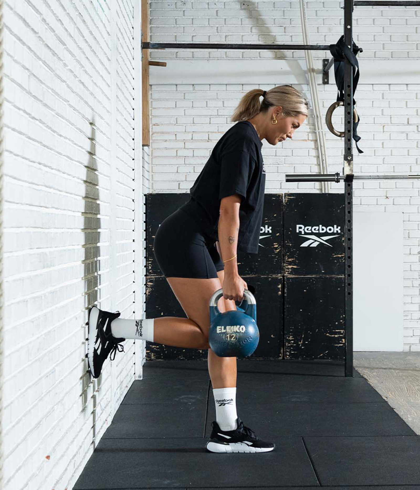A woman performs a single-leg kettlebell exercise with one foot elevated against a wall.