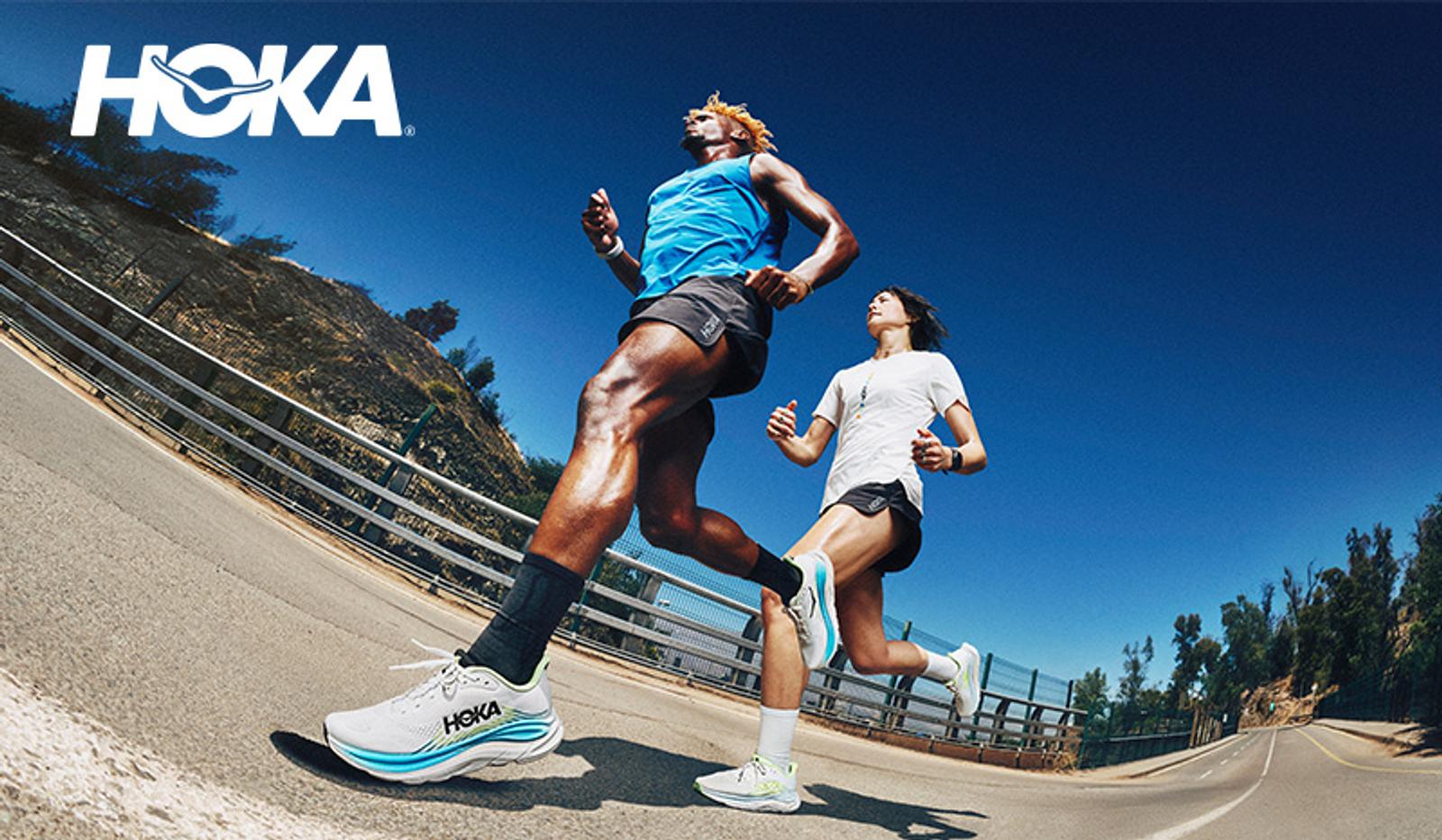 Two runners wearing Hoka shoes run on a sunny outdoor road, with the Hoka logo in the top left.