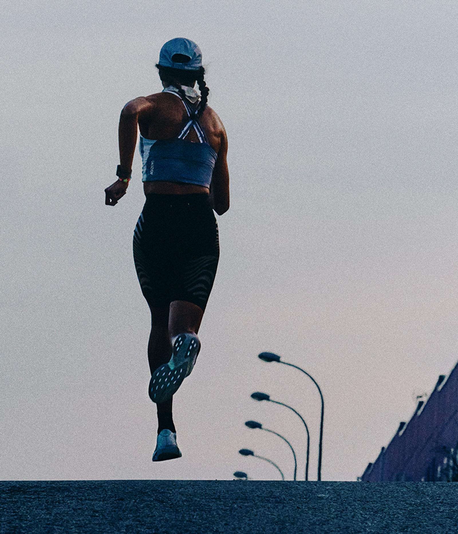 Rear view of a runner in a cap and sports bra, on an incline with streetlights.