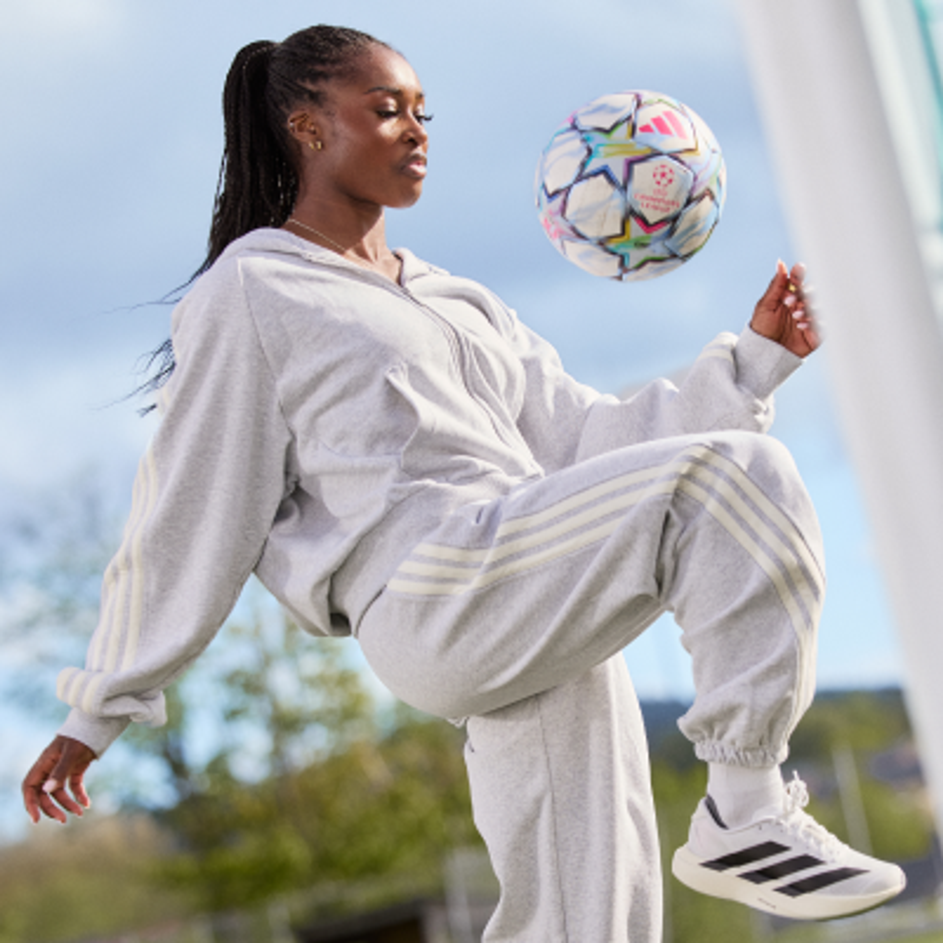 A woman in a grey Adidas tracksuit balancing a colorful soccer ball on her knee.