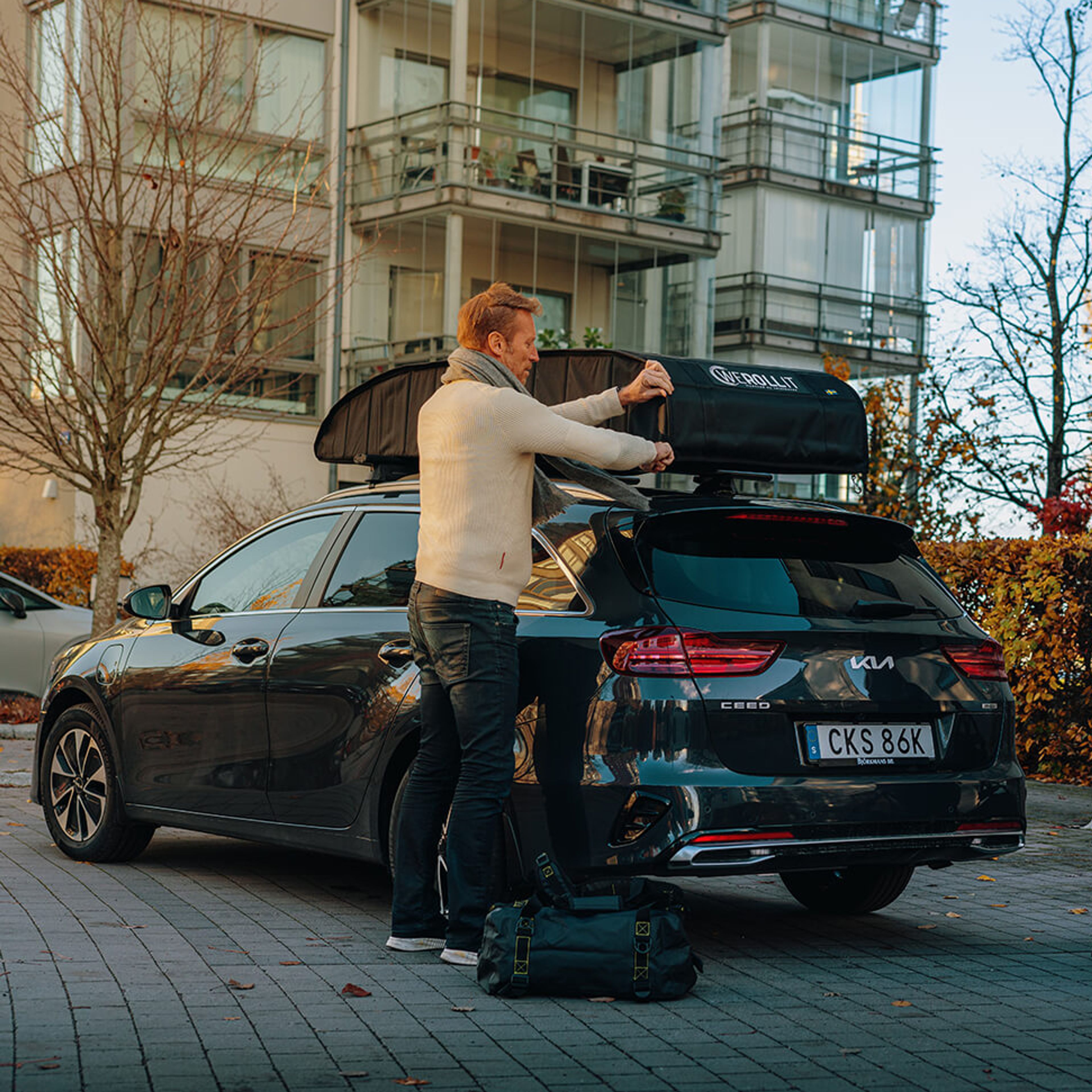 A man loads a soft roof cargo bag onto a black SUV as a woman stands nearby with a duffel bag.