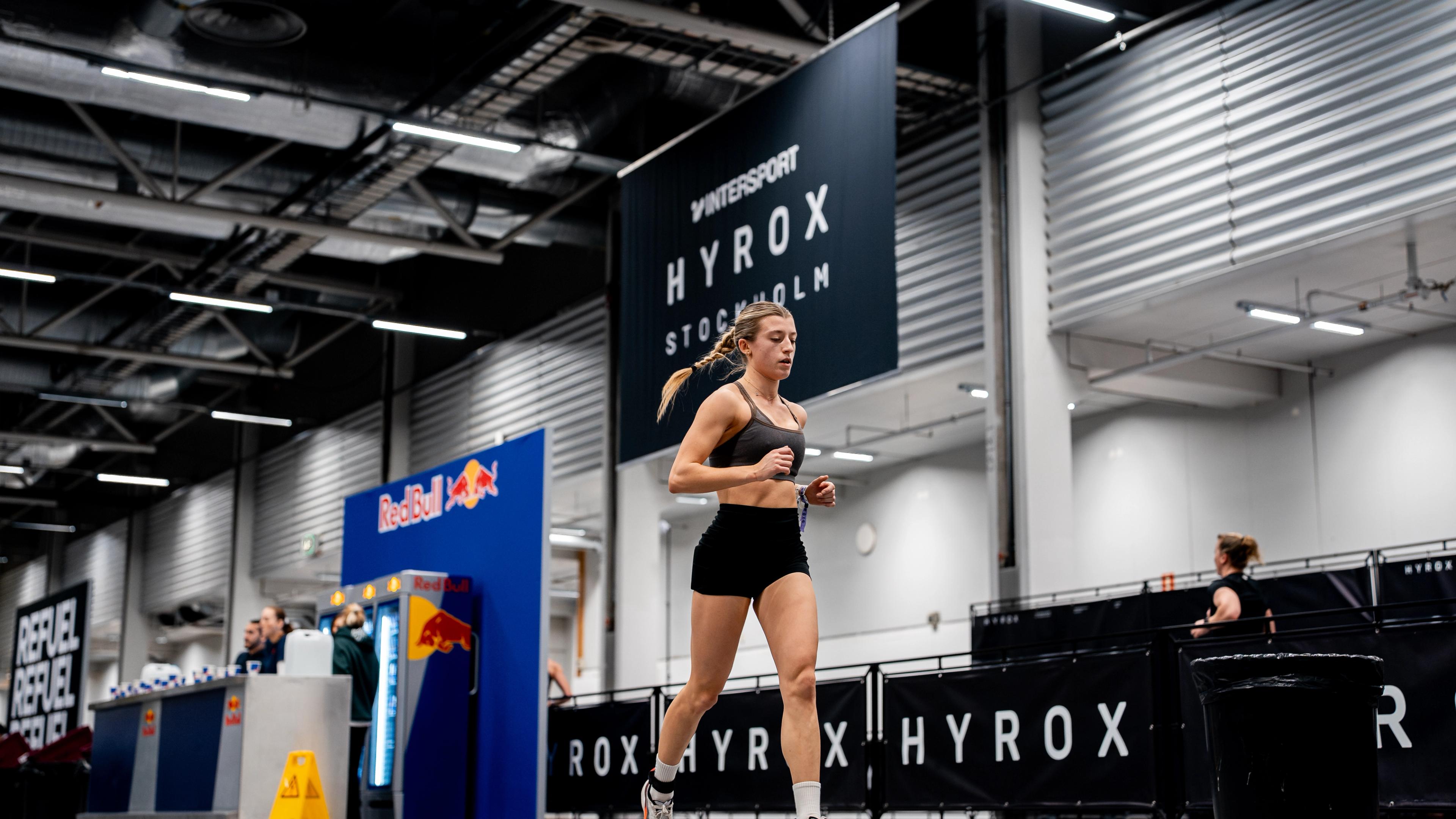 A woman in athletic wear runs indoors at a fitness event, with "HYROX Stockholm" and "Red Bull" branding visible.