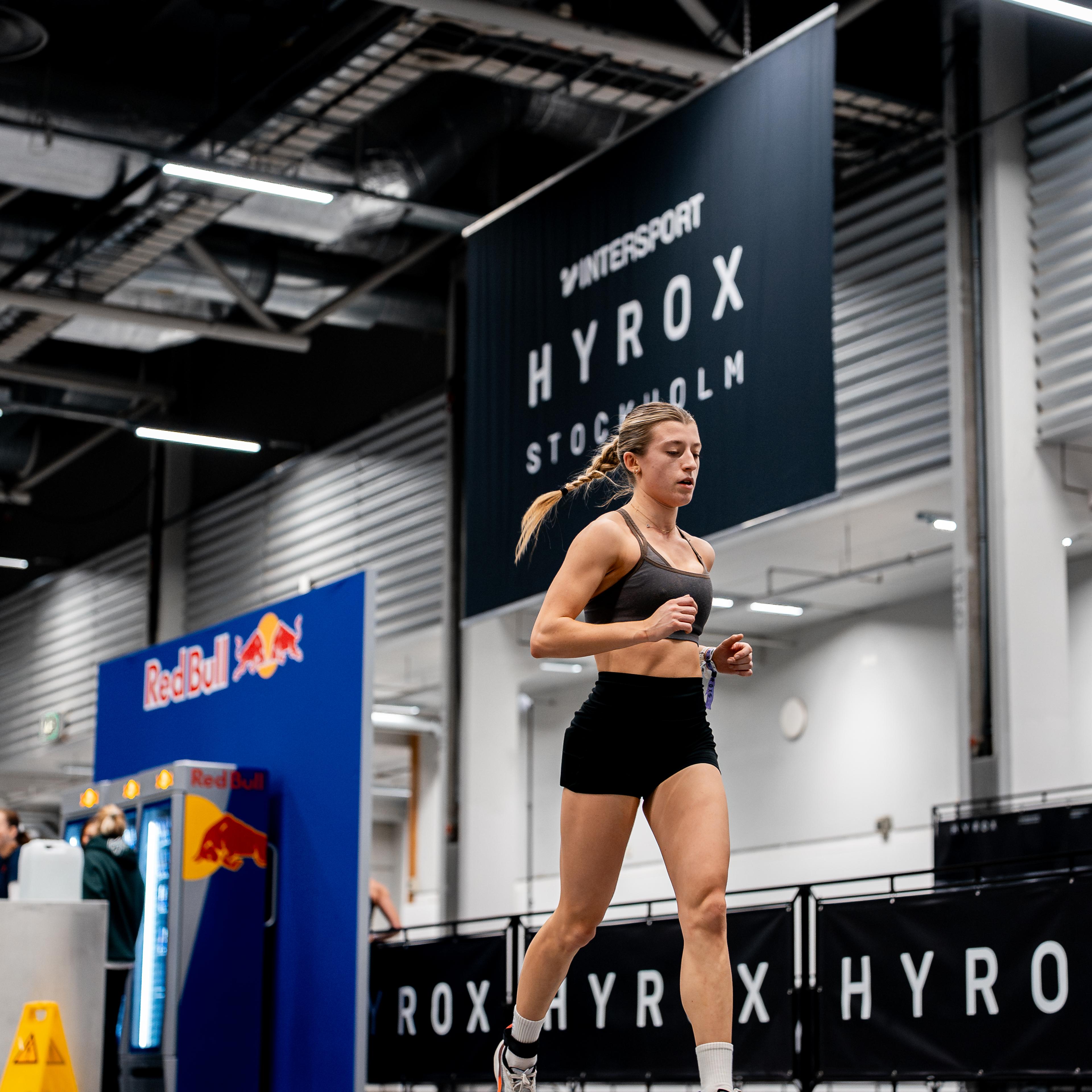 A female athlete runs past a HYROX banner in an indoor arena.