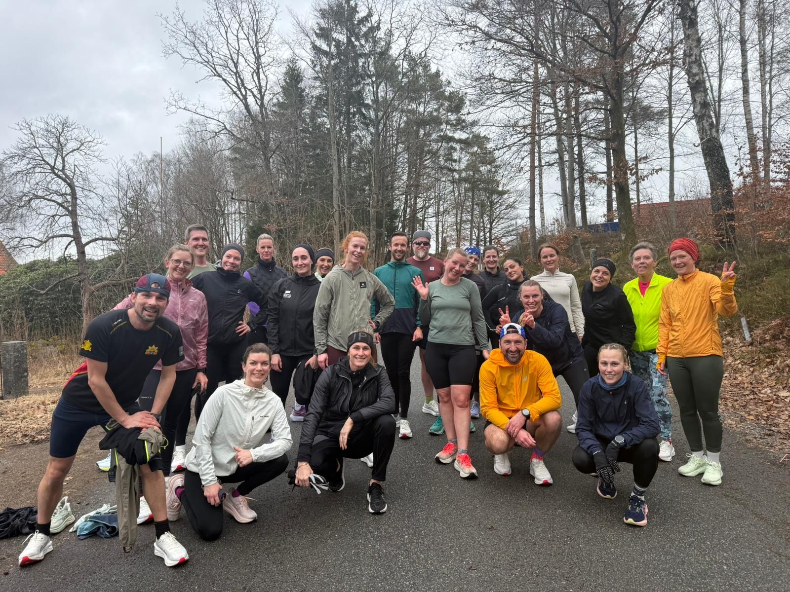 A large group of smiling runners in athletic wear poses on a paved path surrounded by trees.