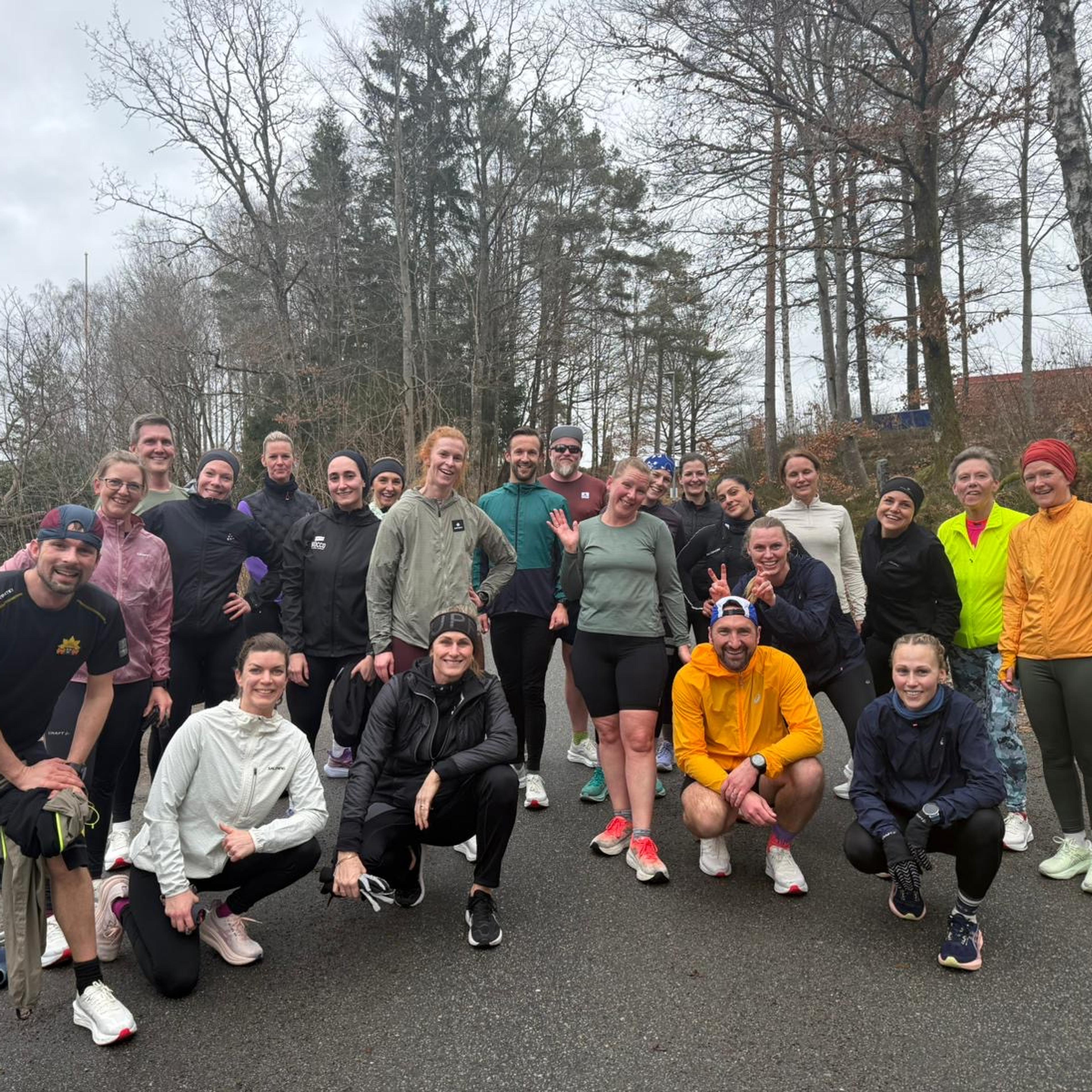 A large group of smiling runners in athletic wear poses on a paved path surrounded by trees.