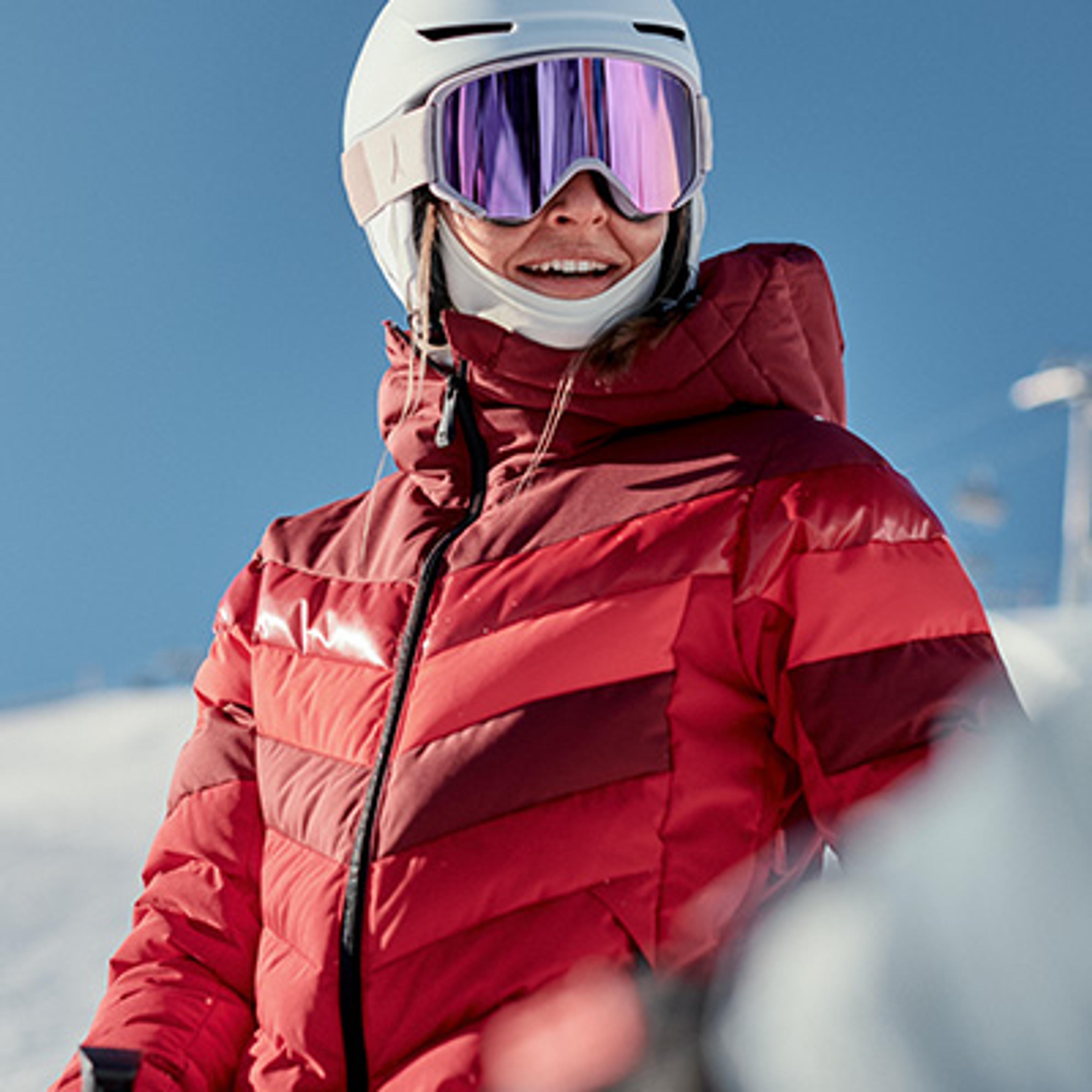 Smiling woman in a white ski helmet, purple goggles, and red quilted ski jacket.