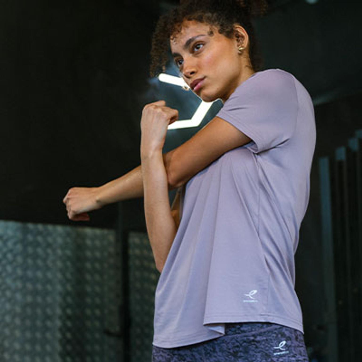 a woman in a purple t-shirt is stretching her arm in a gym .