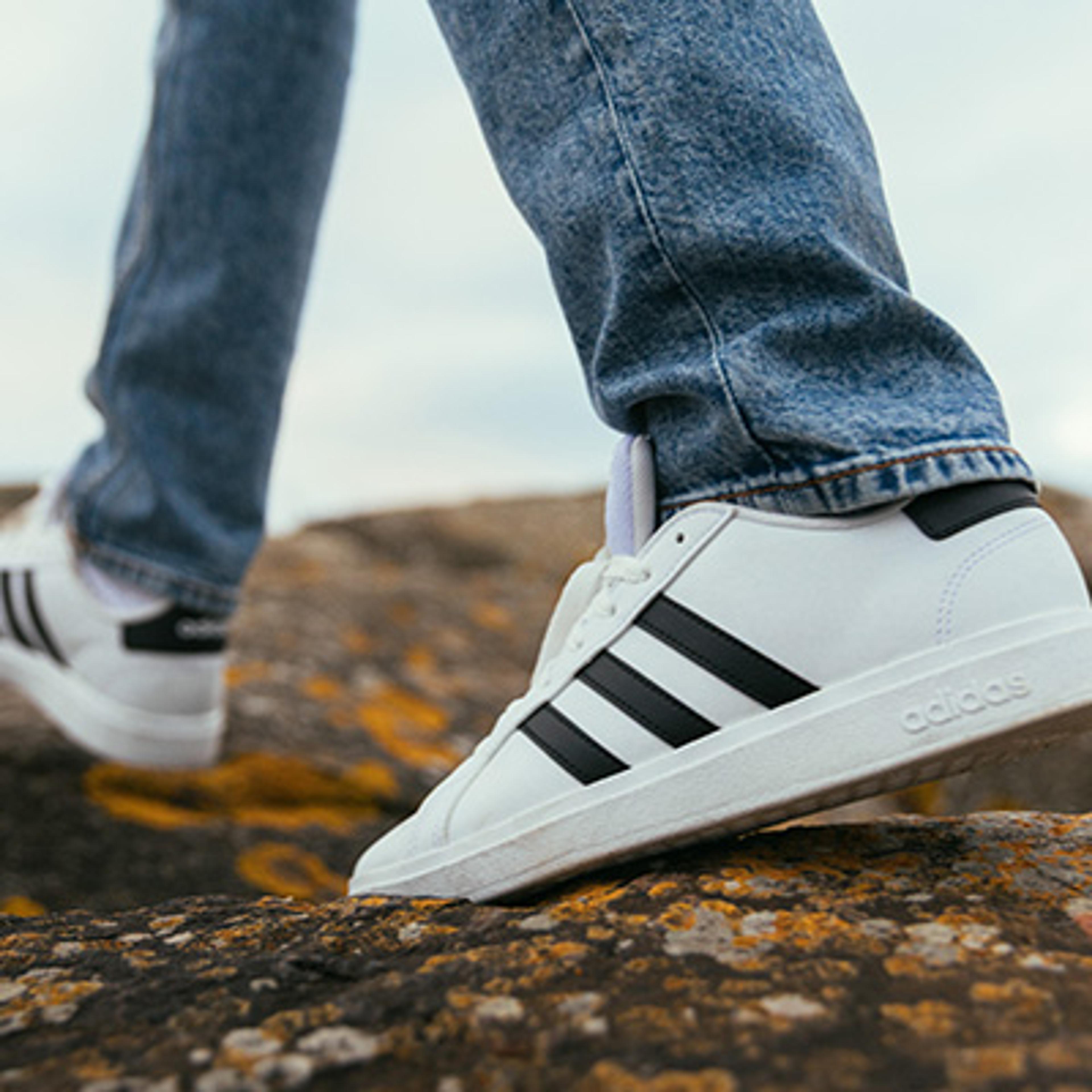 A person in blue jeans and white Adidas sneakers with black stripes walks on a rocky surface.