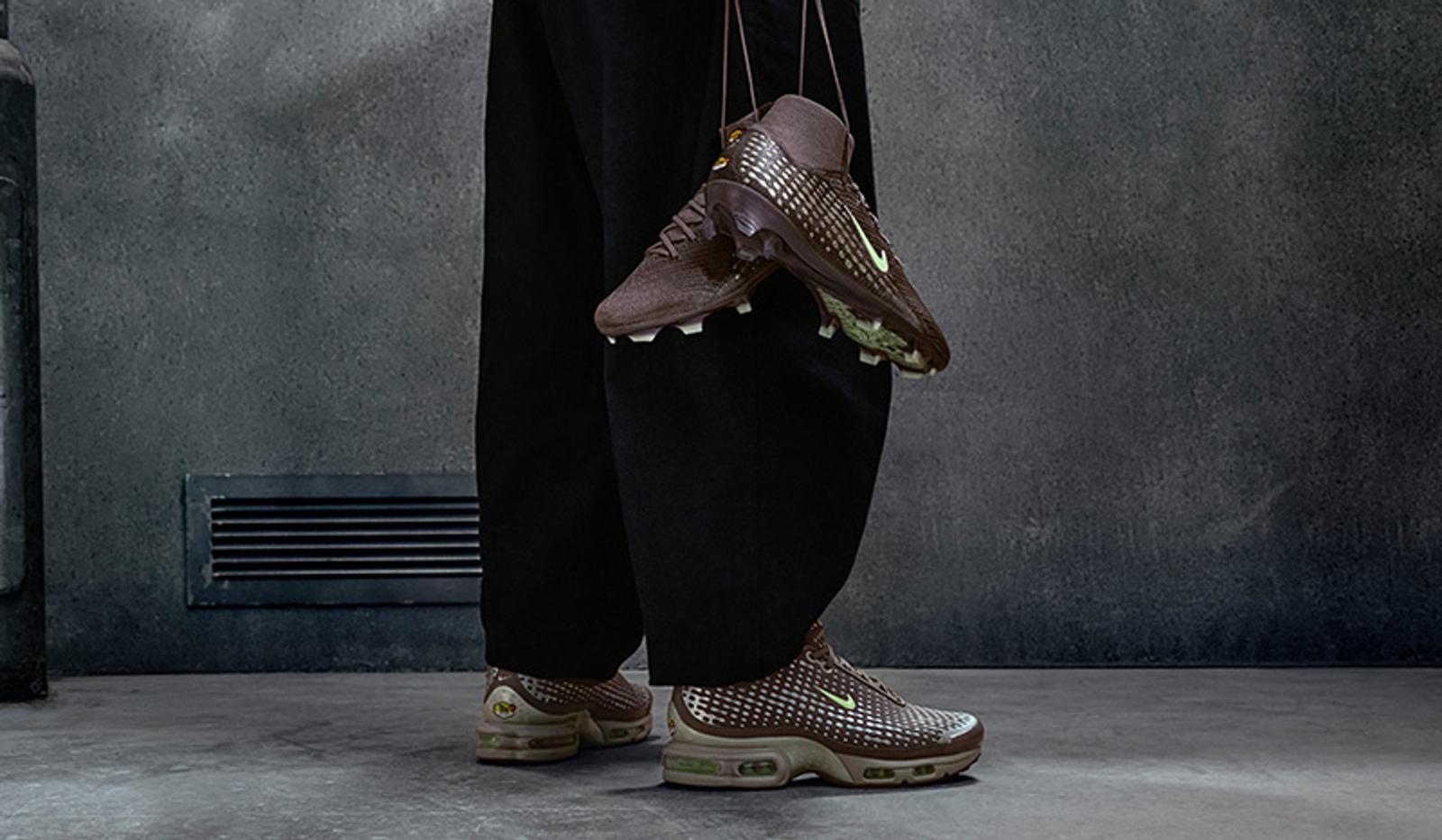 A person wears brown Nike sneakers with a green swoosh and holds a matching pair of brown Nike cleats by strings.
