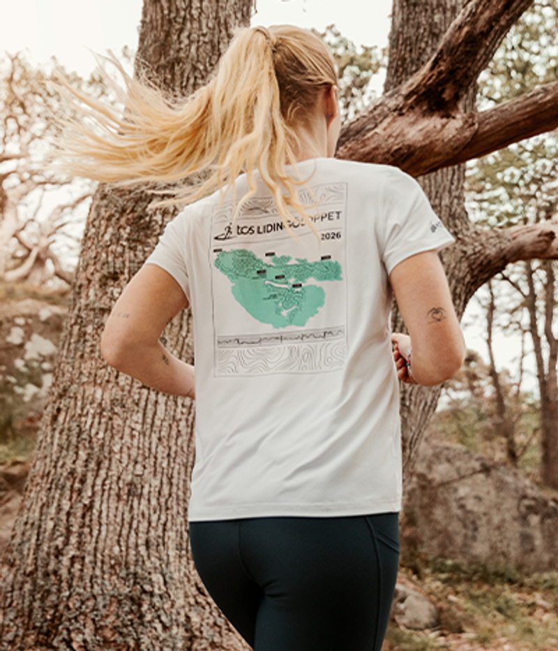 A person with a blonde ponytail runs outdoors, seen from behind, wearing a white t-shirt with a green map graphic and "TCS LIDINGÖLOPPET 2026" on the back.