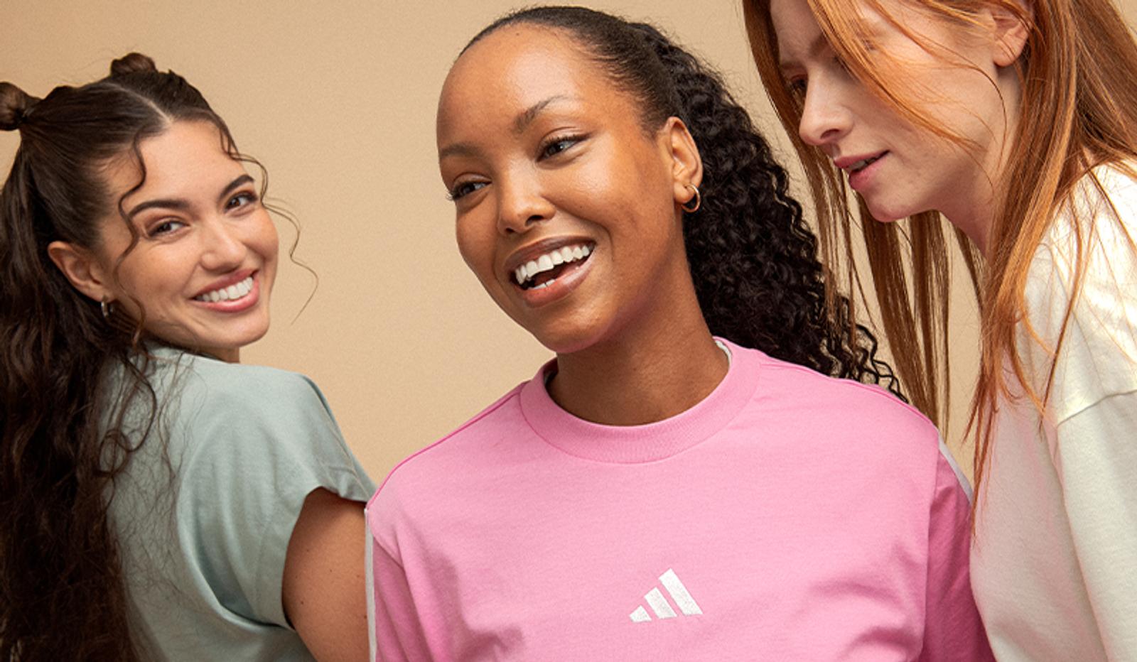 Three diverse women smiling; one wears a pink Adidas t-shirt.